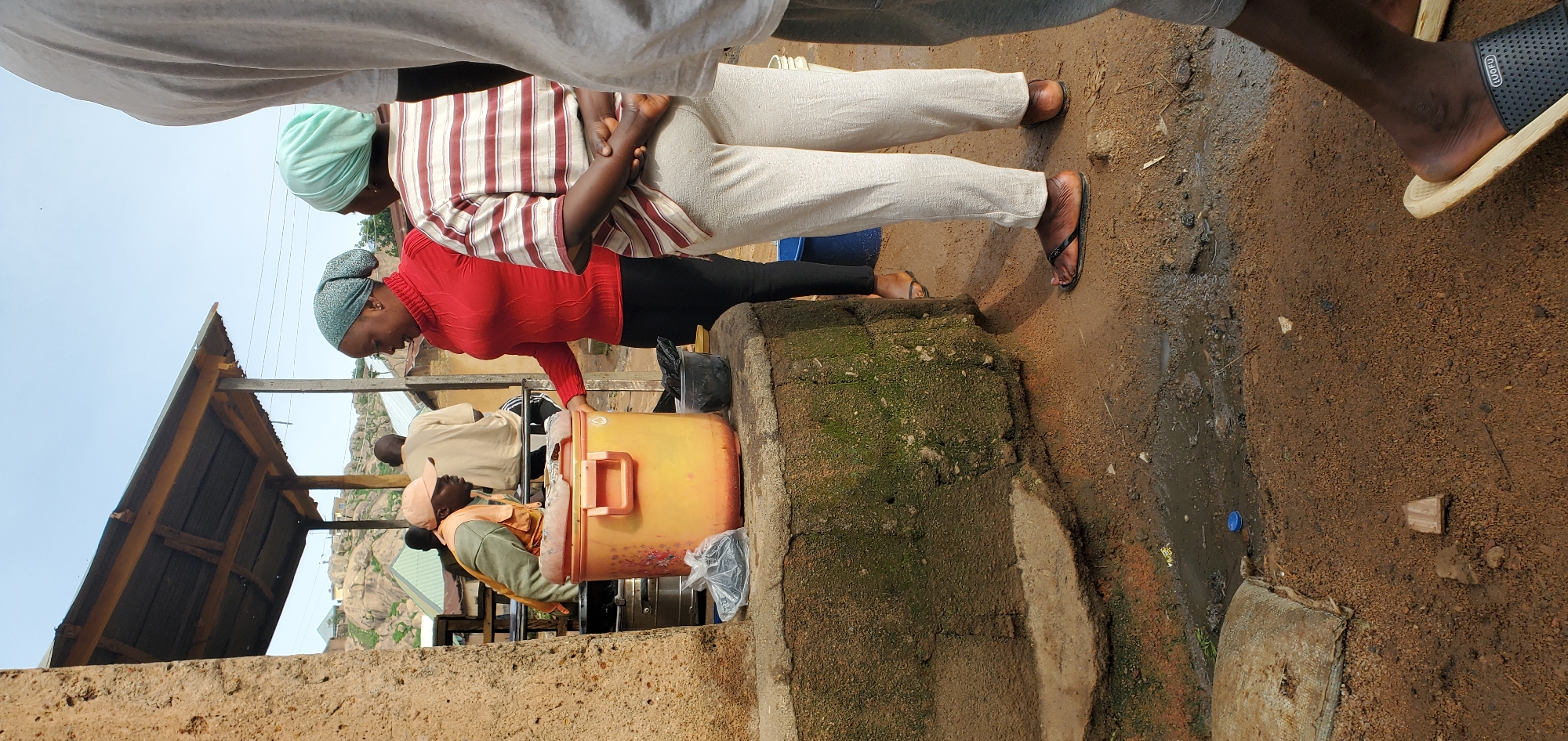 In the city of Unknown, Unknown, a group of individuals are gathered near a rustic outdoor structure with a corrugated metal roof supported by wooden posts. The foreground shows a circular, moss-covered concrete structure with a rough texture, likely a well or a base for a container. On top of this structure, a large, orange, plastic container with a lid is prominently placed. Beside it, a black plastic bag filled with dark items and a smaller black plastic container filled with similar dark items are visible.
Several people are interacting around this area. One individual wearing a red turtleneck sweater and a light blue headscarf is looking towards the orange container, her right hand resting on its lid. Another person, wearing a light blue headscarf and a striped t-shirt, stands with their back to the camera, facing away from the container. To the left, partially obscured, is a person wearing a peach-colored cap and an orange vest over a green shirt, appearing to be engaged with something behind the orange container. Further back, two other individuals are visible; one in a light-colored shirt and the other in a beige garment, both looking away from the foreground.
The ground is a mixture of mud, dirt, and gravel, with a visible puddle of murky water. In the background, a hilly landscape with rocky outcrops and buildings can be seen under a clear sky. The overall scene suggests an informal gathering or a marketplace setting in a rural or underdeveloped urban area.