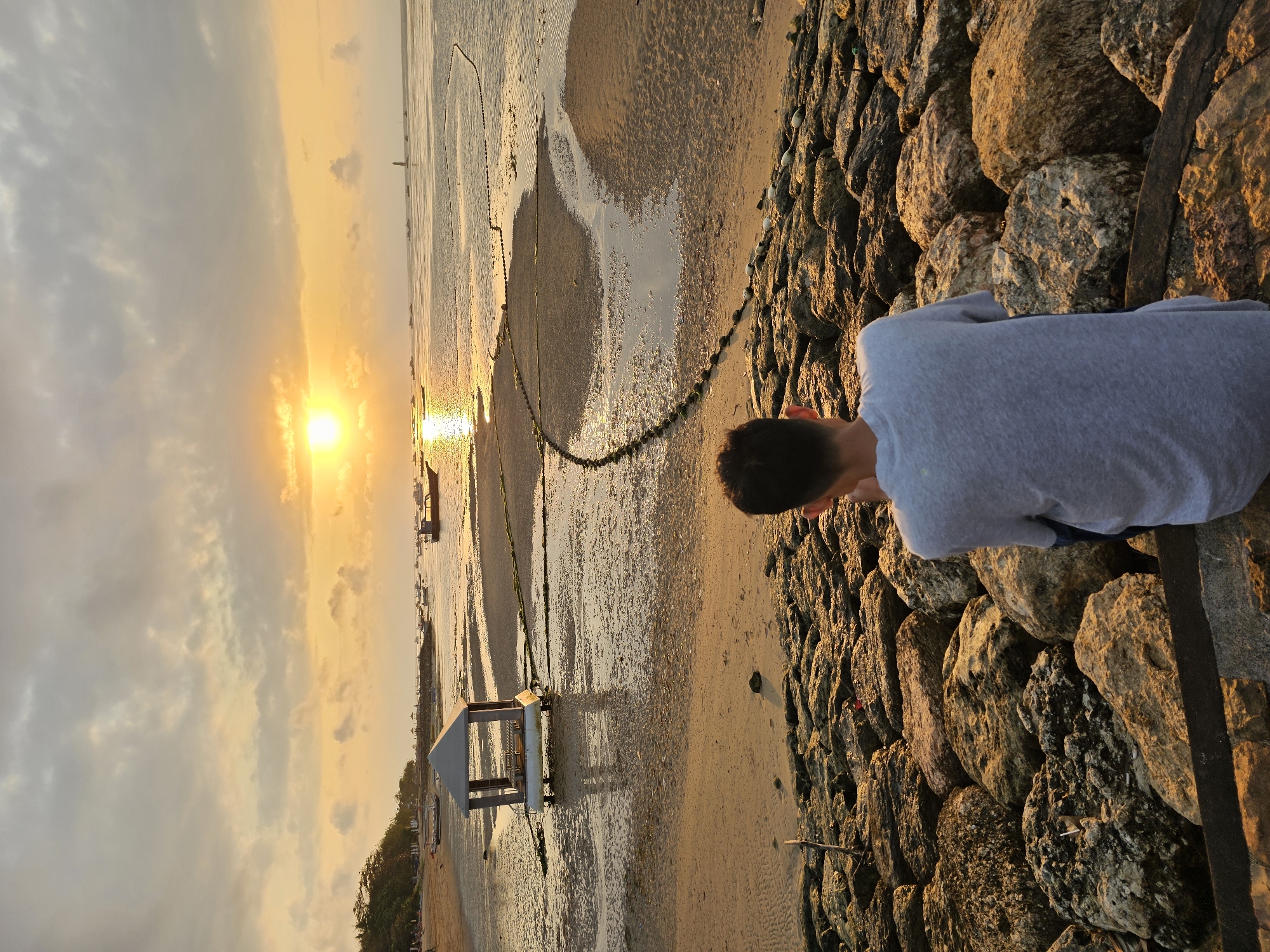 A young person, facing away from the camera, is seated on a large pile of rocks. The person is wearing a gray t-shirt. The setting is a beach at sunset, with the sun low on the horizon and casting a warm glow. The ocean is calm with several small boats visible in the distance. The sand is wet and reflects the light of the sunset. A small, open-sided structure with a pitched roof sits on stilts in the shallow water near the shore. A rope with buoys stretches across the water in front of the structure. The sky is partly cloudy with hues of orange, yellow, and gray. The scene appears to be Sanur Beach in Denpasar, Indonesia, a popular spot for watching the sunrise and sunset.