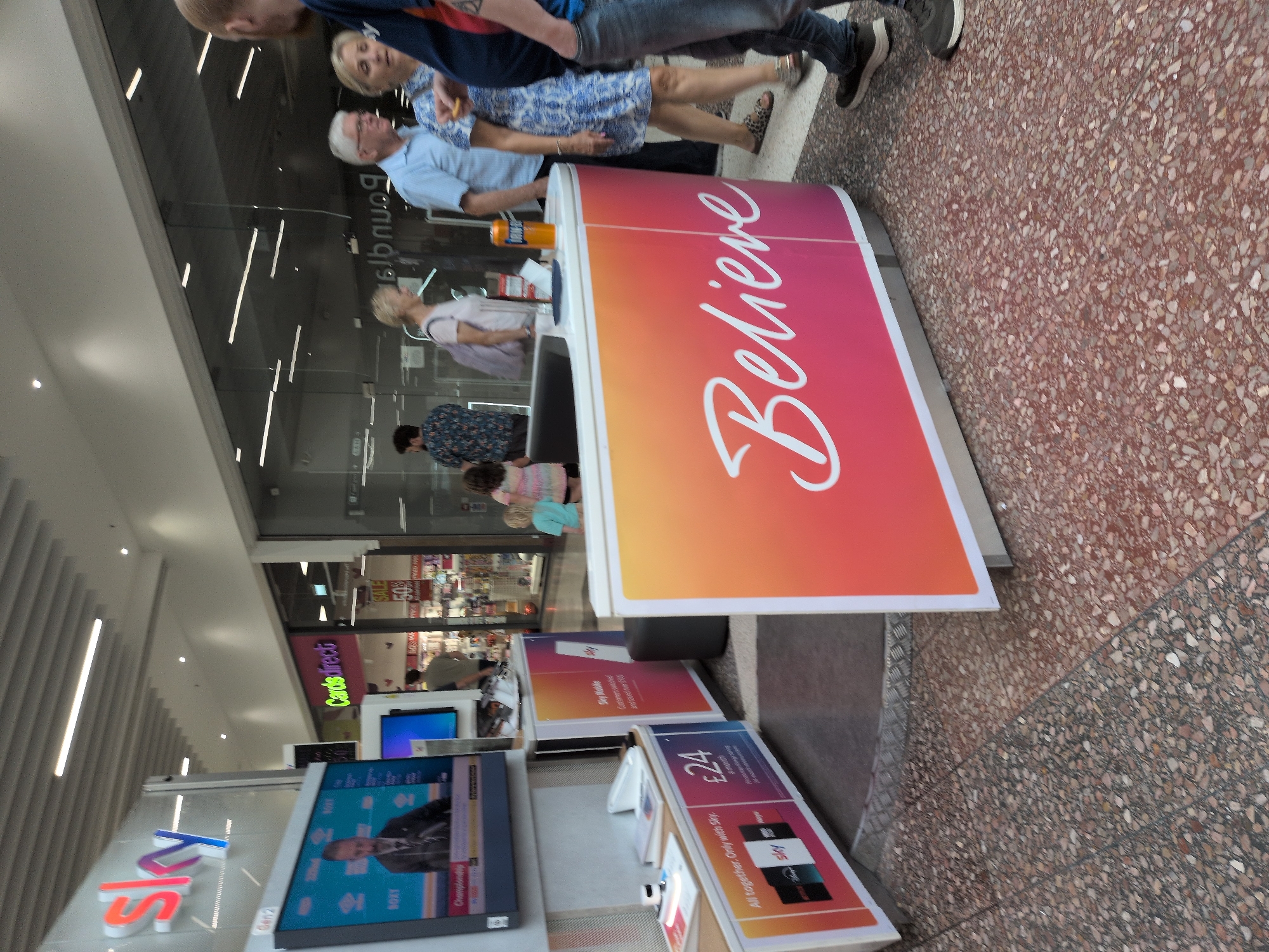The image shows a view inside the Telford shopping centre, United Kingdom. In the foreground, a red and orange promotional stand with the word "Believe" in white cursive script is visible. To the left of the stand, another promotional display featuring Sky products is present, with a screen showing a man in a suit and text that appears to be related to a championship. Several people are scattered throughout the scene. In the mid-ground, a man in a light blue shirt is talking to a woman wearing a blue and white patterned dress. Behind them, other shoppers and a shopfront are visible, including one with a "Cards Direct" sign. The flooring is a speckled terrazzo pattern. The lighting suggests it is daytime, with artificial lights visible on the ceiling.