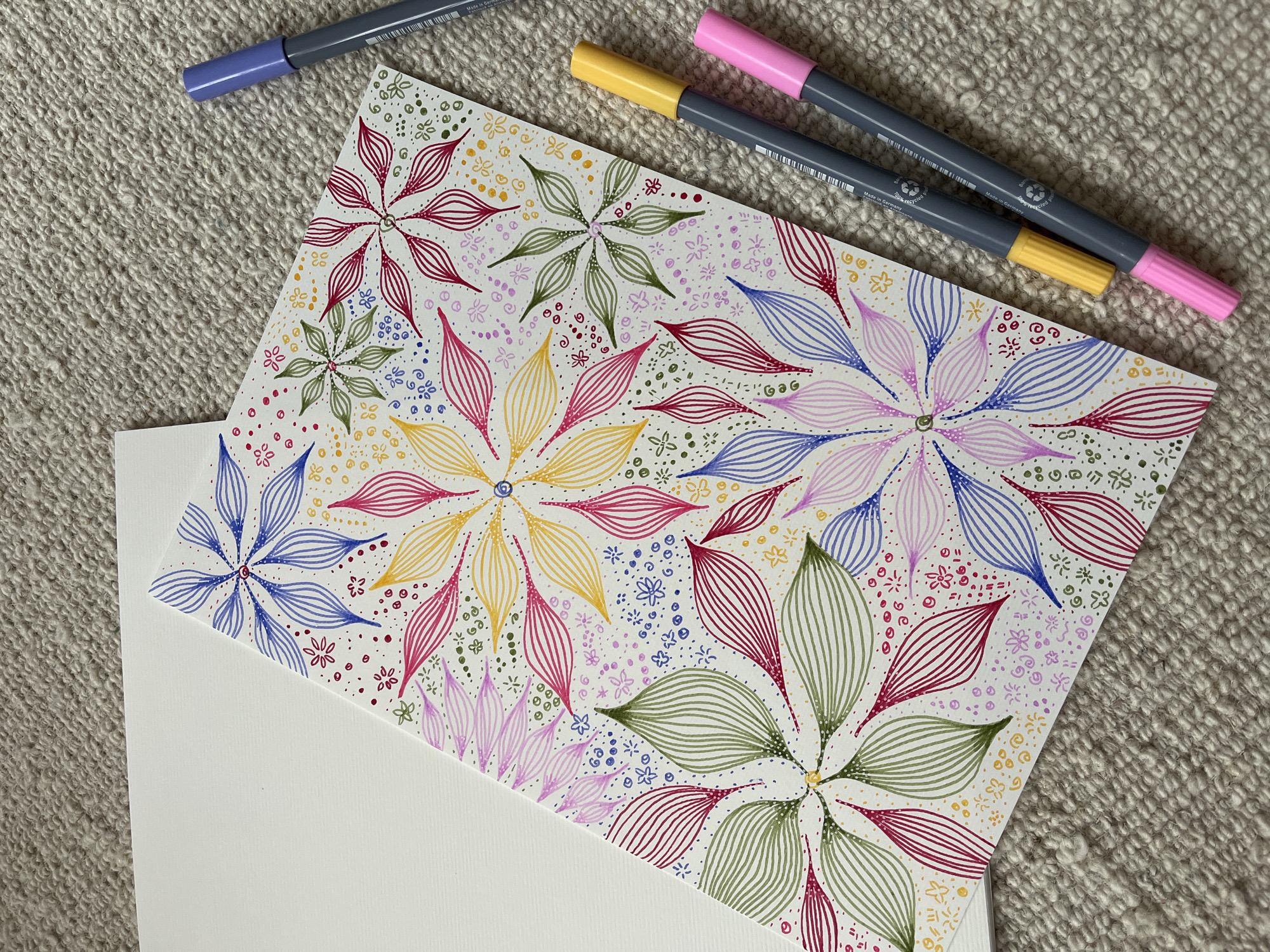 The image shows a close-up view of a drawing featuring colorful, stylized floral designs, created with markers. The drawing is placed on top of a blank white card and resting on a textured beige surface, possibly a rug or fabric. A light purple marker, a yellow marker, and a pink marker are also present. The overall composition is clean and well-lit, suggesting an artistic or crafting setting.