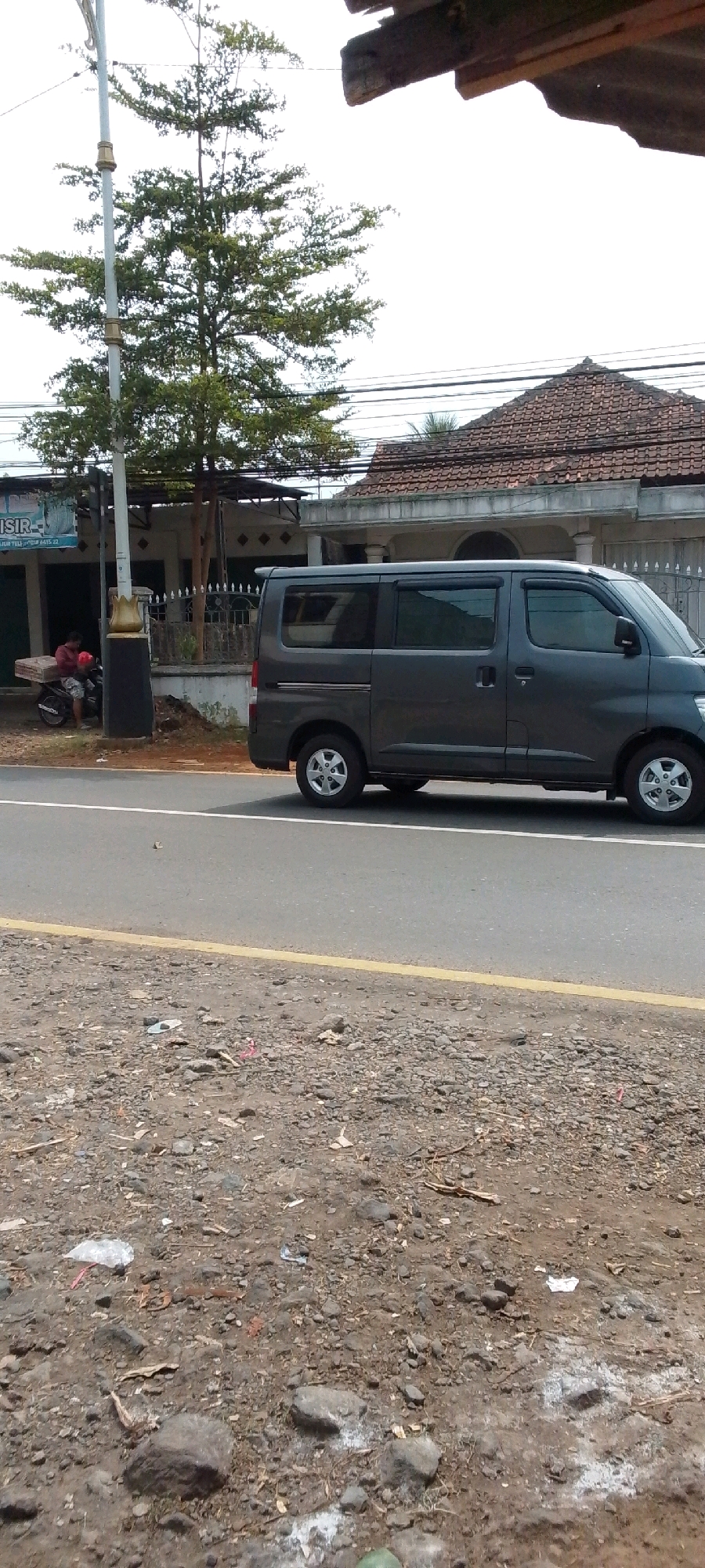 In Cilaku, Indonesia, on a paved road, a dark gray van is parked to the right of a motorcycle. The motorcycle is ridden by a person wearing a red shirt and dark shorts, who is carrying a large, flat object that resembles a mattress or a board on the back of the motorcycle. To the left of the motorcycle and van, a building with a blue sign that reads "SISTEM" is visible. Behind the building, a house with a tiled roof and an open gate can be seen. The scene is framed by a tall, slender tree with green foliage on the left and a wooden overhang on the right, suggesting a roadside location. The ground in the foreground is covered with gravel and debris.