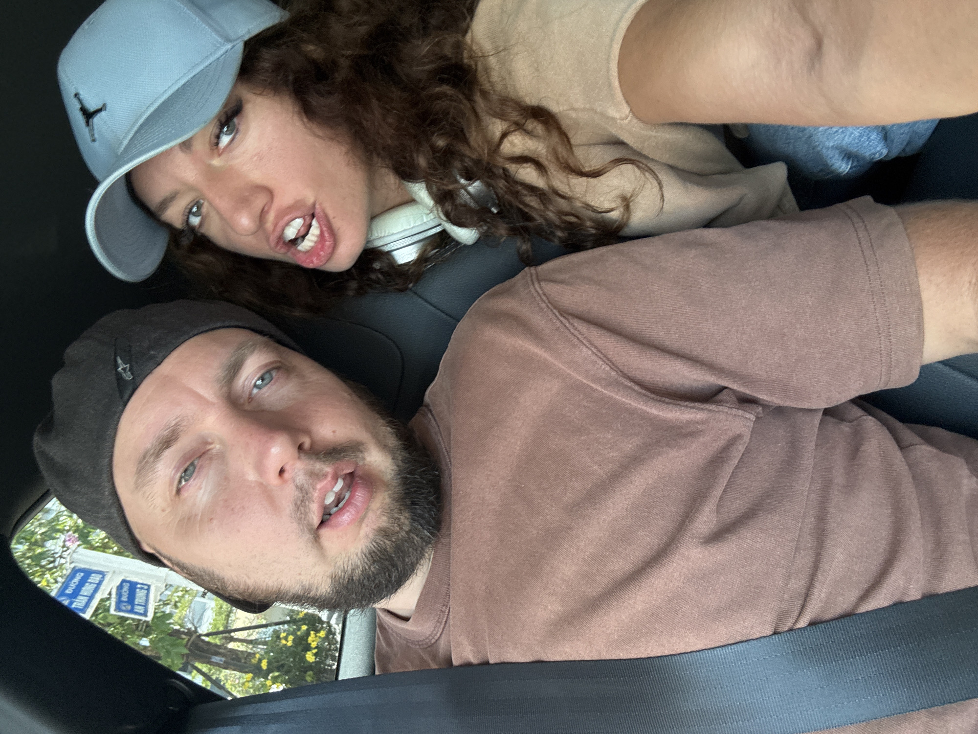 An eye-level, slightly upward-angled photograph captures two individuals inside a vehicle, possibly a car, secured with a visible seatbelt. The foreground features a man with a beard, wearing a dark-colored hat and a reddish-brown t-shirt. His eyes are light, and his mouth is slightly open. Behind him, a woman with curly brown hair is wearing a light blue baseball cap, a beige sweatshirt, and headphones around her neck. She is positioned above the man, partially obscuring the scene behind them. Her mouth is open, revealing her teeth.
Outside the car window, a street sign written in Vietnamese is visible amidst green foliage and a blue sky. The street sign displays the names "DUONG TRAN HUNG DAO" and "DUONG AN TRUNG 3", placing the location in Đà Nẵng, Vietnam. The photograph appears to be taken from inside the car, potentially by one of the occupants.