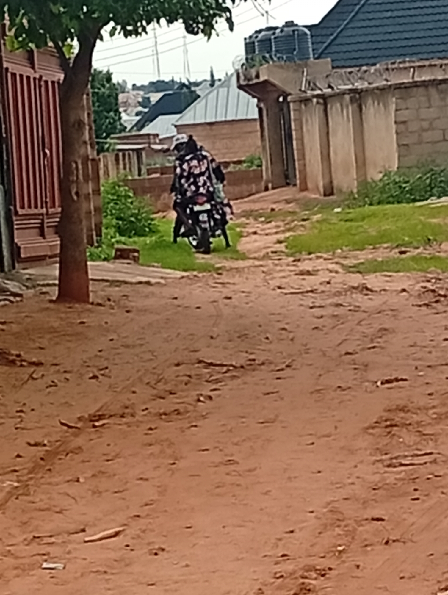 The image depicts a street scene in Tumfure, Nigeria. A person wearing a patterned garment and a white cap rides a motorcycle down a dirt road. The motorcycle is black and white with visible wheels and handlebars. On the left side of the road, there is a red gate and a tree. The background shows buildings with dark roofs, some with water tanks on top, and green grass. The road surface is predominantly reddish-brown dirt with scattered debris. The sky is overcast, suggesting a cloudy day.