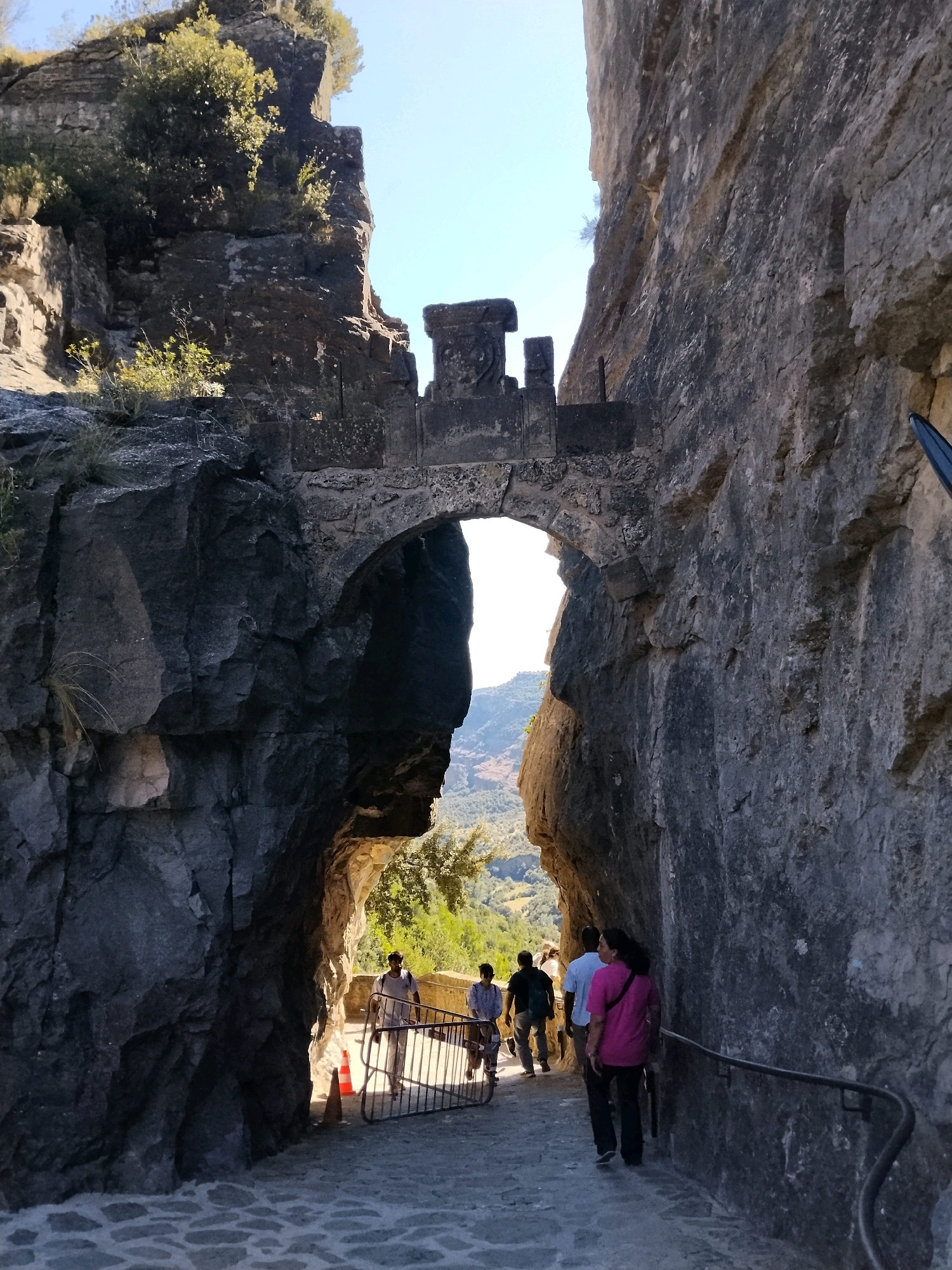 The image shows a narrow passage through a rocky gorge, with an arched stone structure overhead. Several people are walking through the passage, some from behind. The path is paved with cobblestones. The scene appears to be in Sant Quirze Safaja, Spain, likely at the entrance to the Sant Miquel del Fai monastery complex. The architecture suggests historical significance, with the rough-hewn rock walls contrasting with the carved stone of the arch. The lighting indicates it is daytime with clear skies.