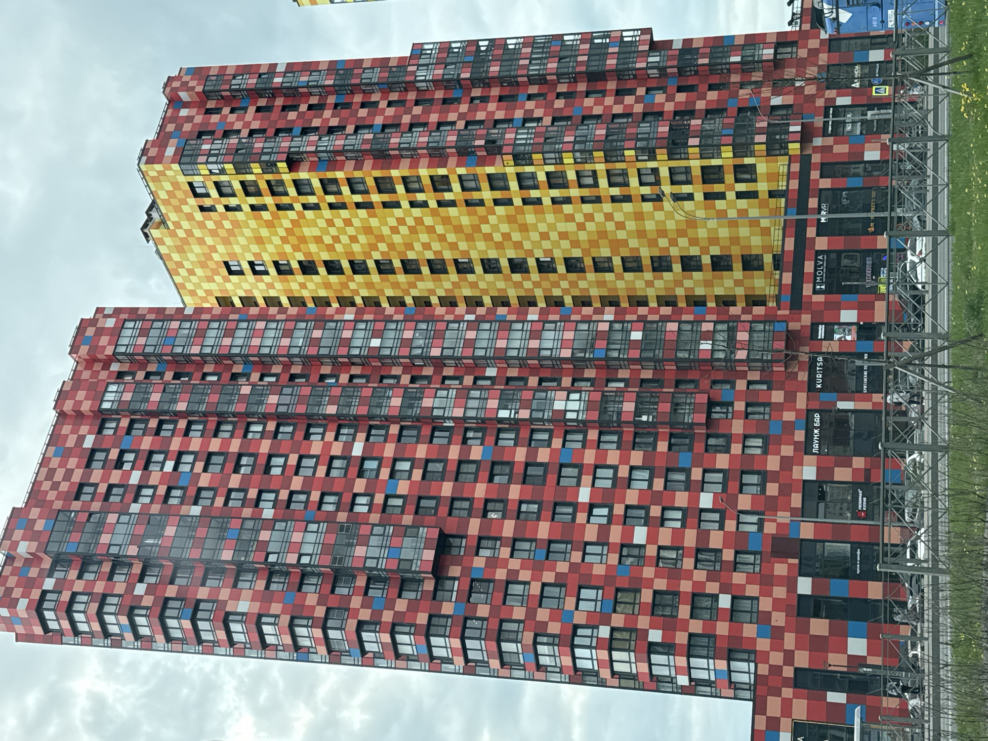 A low-angle, medium shot depicts two tall, angular buildings with a mosaic-like facade. The building in the foreground is primarily red with small squares of pink, blue, and white interspersed throughout, while the building behind it is yellow. Both buildings feature many square windows across all floors. The base of the building in the foreground is lined with storefronts. Russian text is visible on the storefront signs, and the English word "MOLVA" is prominent on one of the signs. A metal fence and green vegetation are visible on the right side of the image. The sky is overcast. The location is Saint Petersburg, Russia.