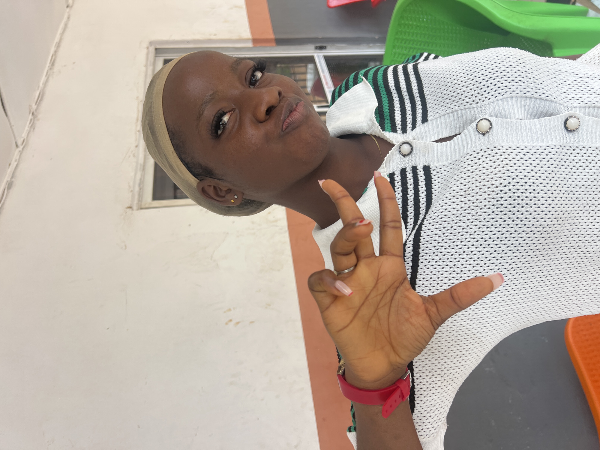 A young Black woman wearing a beige wig cap and a white, black, and green striped polo shirt is shown. She is making a peace sign with her right hand, which has manicured nails. A red watch is visible on her left wrist. She is indoors, with a white and orange wall behind her. Green and red chairs are also visible in the background. The scene appears to be in Nkpor, Nigeria.
