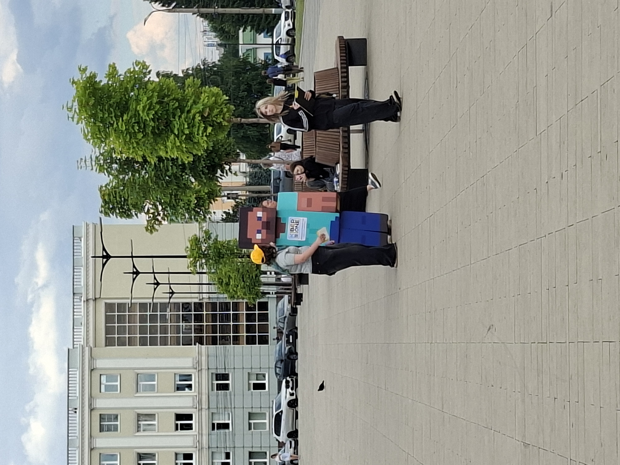 The image depicts an outdoor scene in Izhevsk, Russia, featuring several people and a large, pixelated costume resembling a Minecraft character. Two women are seated on a park bench. One, with blonde hair, is leaning back and holding a book or tablet. The other, with darker hair, is seated closer to the foreground, also appearing to be holding something. In front of the bench, two individuals are interacting with the Minecraft costume. One person, wearing a yellow cap, is standing and appears to be talking to or handing something to the person inside the costume. The costume is predominantly teal and pink, with a square head and body. A sign is visible on the costume with text that reads "ВПР ИОНЕ".
The setting appears to be a public space with a paved walkway, trees, and a large building in the background. The building has a modern architectural style with many windows. Cars are parked along the street in the background, suggesting an urban environment. The sky is cloudy, indicating a possibly overcast day. The overall mood seems casual and perhaps related to an event or promotional activity involving the costume. The time of day appears to be daytime, given the lighting conditions.