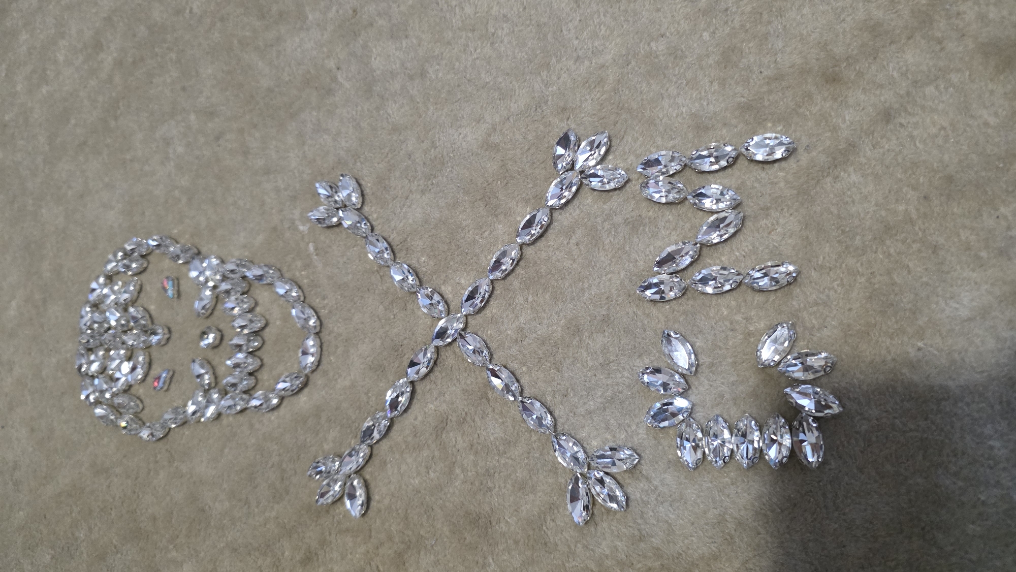 The image displays a detailed, top-down view of numerous clear, faceted, marquise-shaped rhinestones arranged on a soft, light beige or tan-colored fabric surface, possibly felt or velvet. No people are visible in the scene.
The rhinestones are meticulously arranged to form distinct symbols and characters. On the far left, a circular or oval shape is formed by a continuous chain of rhinestones, enclosing several smaller, scattered rhinestones within its boundary. These inner stones catch light and exhibit subtle colorful reflections, including hints of pink and blue.
In the center of the frame, a large and prominent "X" symbol is created by two intersecting lines of rhinestones. Each line is composed of several marquise stones laid end-to-end, with additional stones fanning out from the ends of the diagonals, adding to its decorative quality.
To the right of the "X", the number "2" is clearly formed by arranging the rhinestones. Below the "2", another stylized curved shape, resembling a cursive "S" or an ornate flourish, is created with a line of rhinestones, featuring additional stones at its terminals.
The overall scene suggests a decorative crafting project or an artistic display. The reflective qualities of the rhinestones are highlighted by soft, even lighting, which does not cast harsh shadows. The texture of the background fabric is visible, providing a contrasting matte surface to the sparkling jewels.