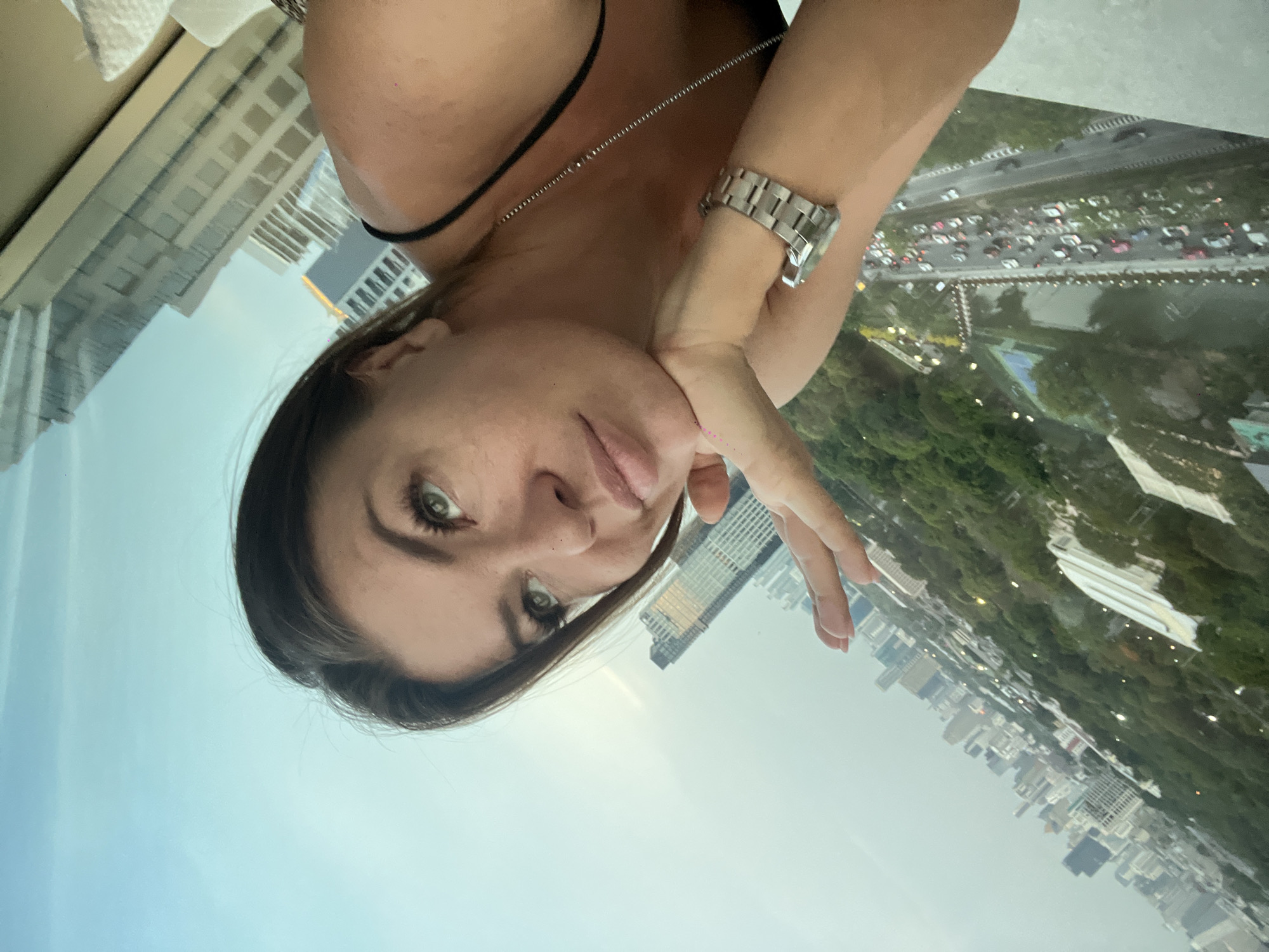 A woman poses in front of a window overlooking Bangkok, Thailand. Her left cheek rests on her left hand and she looks directly at the camera. She is wearing a black tank top and a silver watch. A thin silver necklace rests on her upper chest. Outside the window is a view of a busy city street, lush greenery, and a cityscape of skyscrapers. Heavy traffic fills the streets of Bangkok.