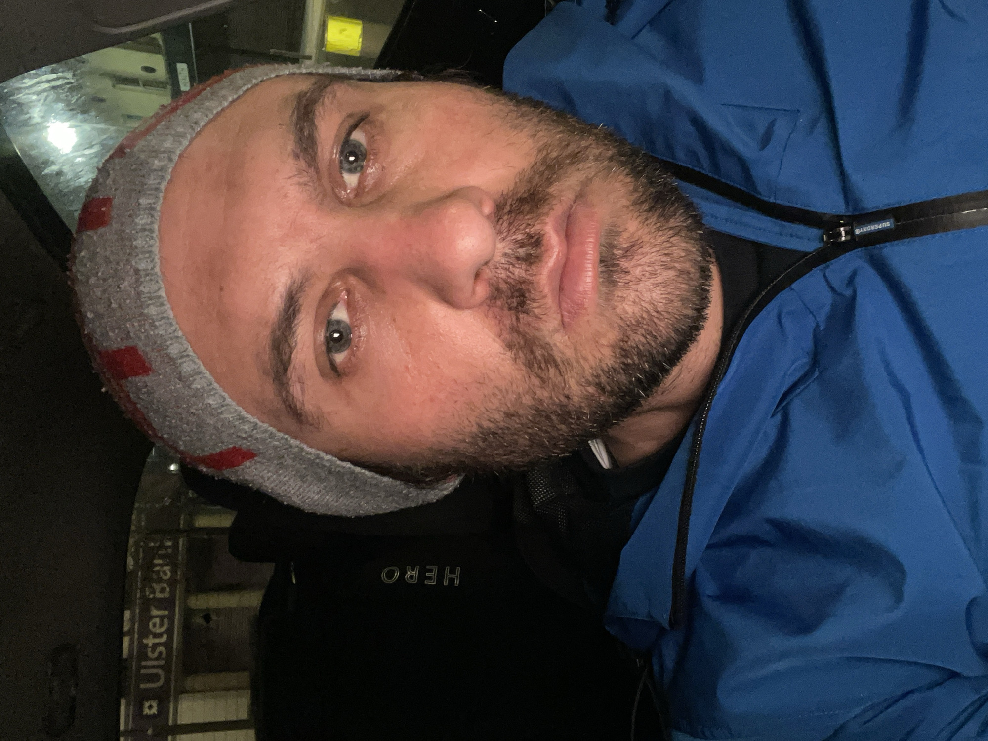 A man is in a car near an Ulster Bank in Newry, United Kingdom. He is wearing a gray beanie with red stripes, a black shirt with the word "HERO" on it, and a blue jacket labeled "Superdry". The time is likely nighttime.