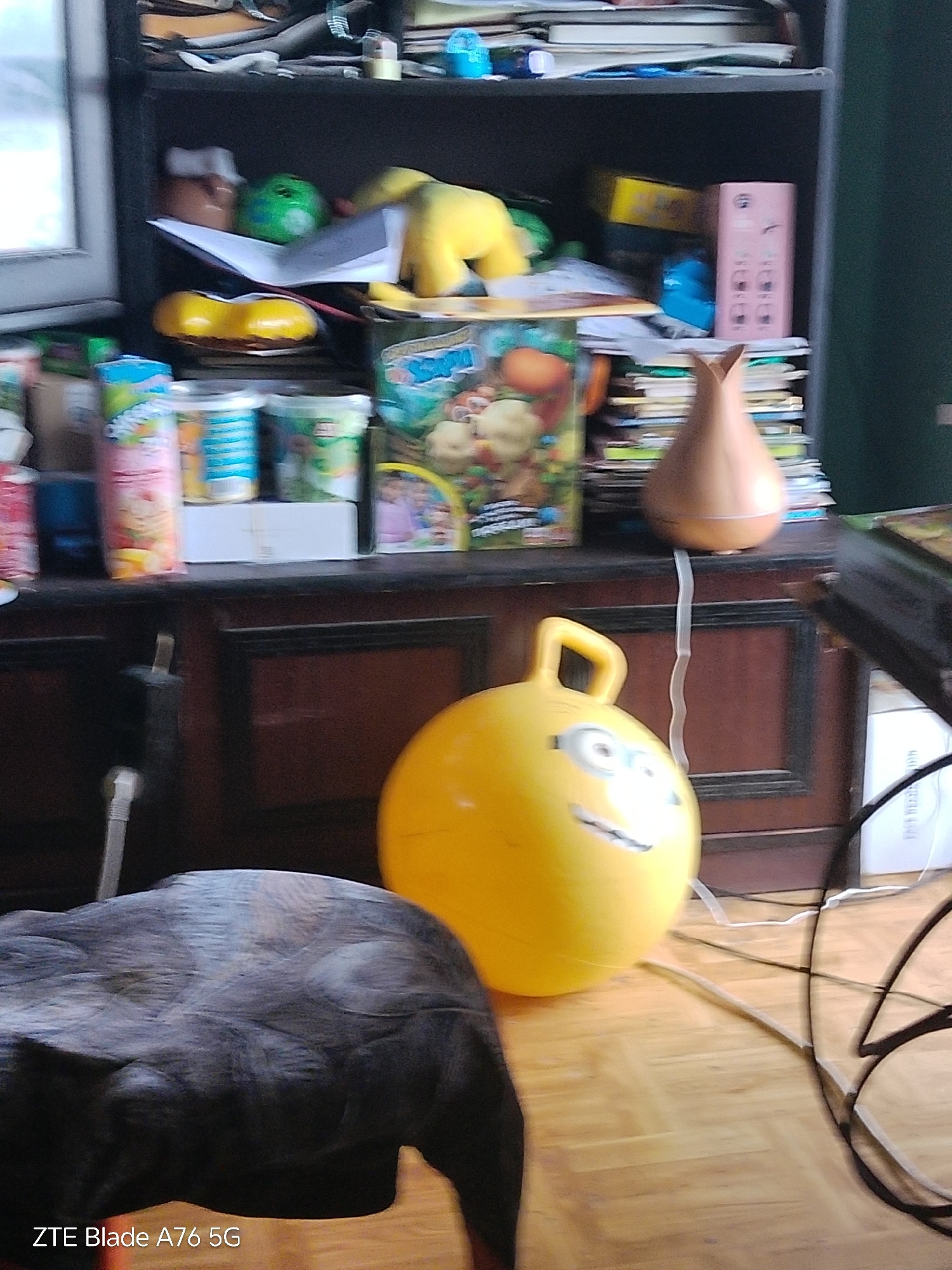 The scene is an interior room, presumed to be in the city of Stara Zagora, Bulgaria. The image depicts a cluttered domestic environment, devoid of people.
In the immediate foreground, to the lower-left, a large, dark, textured textile object, possibly a pet bed or cushion, occupies the space. The lower-left corner bears the watermark "ZTE Blade A76 5G".
Centered in the mid-ground is a bright yellow, inflatable space hopper featuring a handle and a cartoon character face, specifically resembling a Minion, resting on a light-colored wooden parquet floor. To its right, a cluster of black and white electrical cables lies on the floor, partially obscuring a black, open-framed structure.
The background features a two-tiered dark wooden furniture unit. The lower section is a cabinet or chest with two visible paneled doors. On its surface, from left to right, are: various children's product containers including a pink bottle with a partial "Shopkins" label, several smaller plastic tubs, and a colorful box depicting fruit and characters, likely a board game or toy set. Behind these, a white cardboard box is situated. To the right, a light brown, tear-drop-shaped essential oil diffuser is visible, with a white power cord extending downwards from its base.
The upper section is a shelving unit or bookcase. Its shelves contain numerous items: the top shelf holds stacks of books and papers along with small blue and brown objects. The middle shelf displays a brown container, a green spherical object, more papers, a yellow box, a blue object, and a pink rectangular object resembling a decorative box or device holder, interspersed with additional books and papers. A partially visible window providing natural light is on the far left.