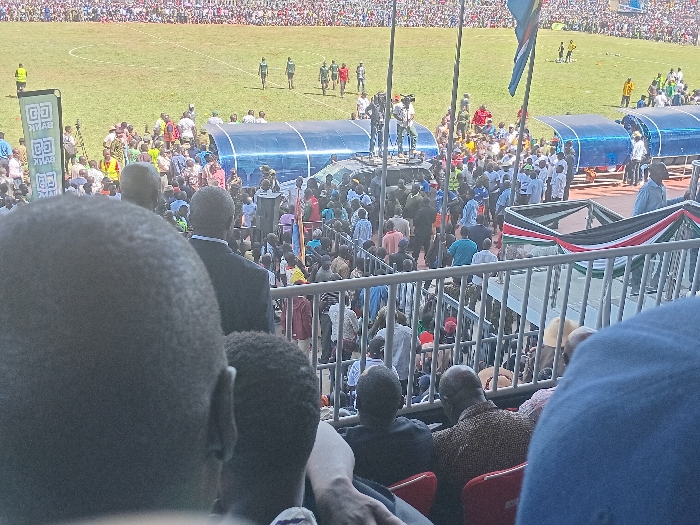 This image depicts a large public gathering within a stadium, likely in Siaya, Kenya, under bright daytime conditions. The photograph is taken from an elevated position within the stands, looking down at the field and a dense crowd of people.
In the foreground, the backs of several individuals' heads are visible, some seated in red stadium chairs. Beyond them, a metal railing separates the viewing area from a lower section packed with spectators. On the grass field, which appears to be a sports pitch, several people are present, including some dressed in green and red sports attire. Two blue, tunnel-like structures, possibly portable dugouts, are situated near the edge of the field.
A prominent raised platform or stage is set up closer to the stands, around which a large congregation of people has gathered. Individuals, including what appears to be security personnel or officials, are standing on this platform. A large banner with the word "BANK" is visible on the field, suggesting sponsorship or a commercial presence.
The Kenyan national flag, characterized by its black, red, and green stripes, is prominently displayed on a barrier in the mid-ground. Another flagpole nearby carries a flag with blue, yellow, green, and red colors. The bright sunlight suggests clear weather. The sheer number of attendees and the presence of national symbols indicate a significant public event, potentially a political rally, a major sporting event, or a large community celebration. The scene is bustling with activity, reflecting a lively and engaged crowd.