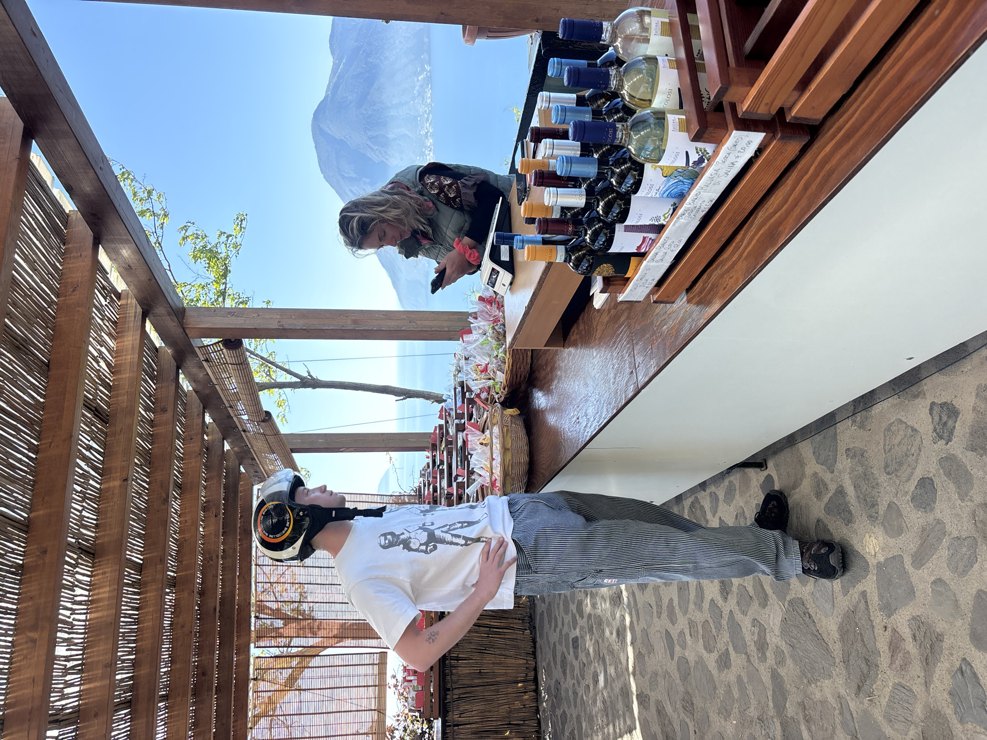 The image captures an outdoor wine shop or tasting area, likely on an island, under a clear blue sky. In the foreground, a man stands, appearing to look upwards, wearing a white t-shirt with a graphic of an astronaut or robot, striped pants, and a white motorcycle helmet with "COOL LINE" visible. His left hand rests on his hip.
Behind him, a wooden counter displays numerous wine bottles, predominantly from the "COLOSI" brand, with some labels also indicating "SALINA." Small signs on the counter provide details and prices, such as "SALINA BIANCO CARODI," "MALVASIA SECCA (DRIED) DI SALINA 20,00," and "SALINA ROSSO 15.00." Baskets filled with wrapped goods are also visible on the counter.
A woman stands behind the counter, dressed in a jacket, intently looking at her phone. Her light-colored hair is tied back with a red scrunchie, and the letters "SAR" are faintly visible, possibly part of "SARAH" on a hair accessory.
The setting features an open wooden pergola structure with slatted bamboo-like screens, casting patterned shadows on the stone-paved ground. In the background, a vast body of water stretches towards distant, rugged mountains or hills, strongly suggesting a scenic coastal or volcanic island landscape, consistent with Lipari, Italy. The bright sunlight and clear blue sky indicate it is daytime.