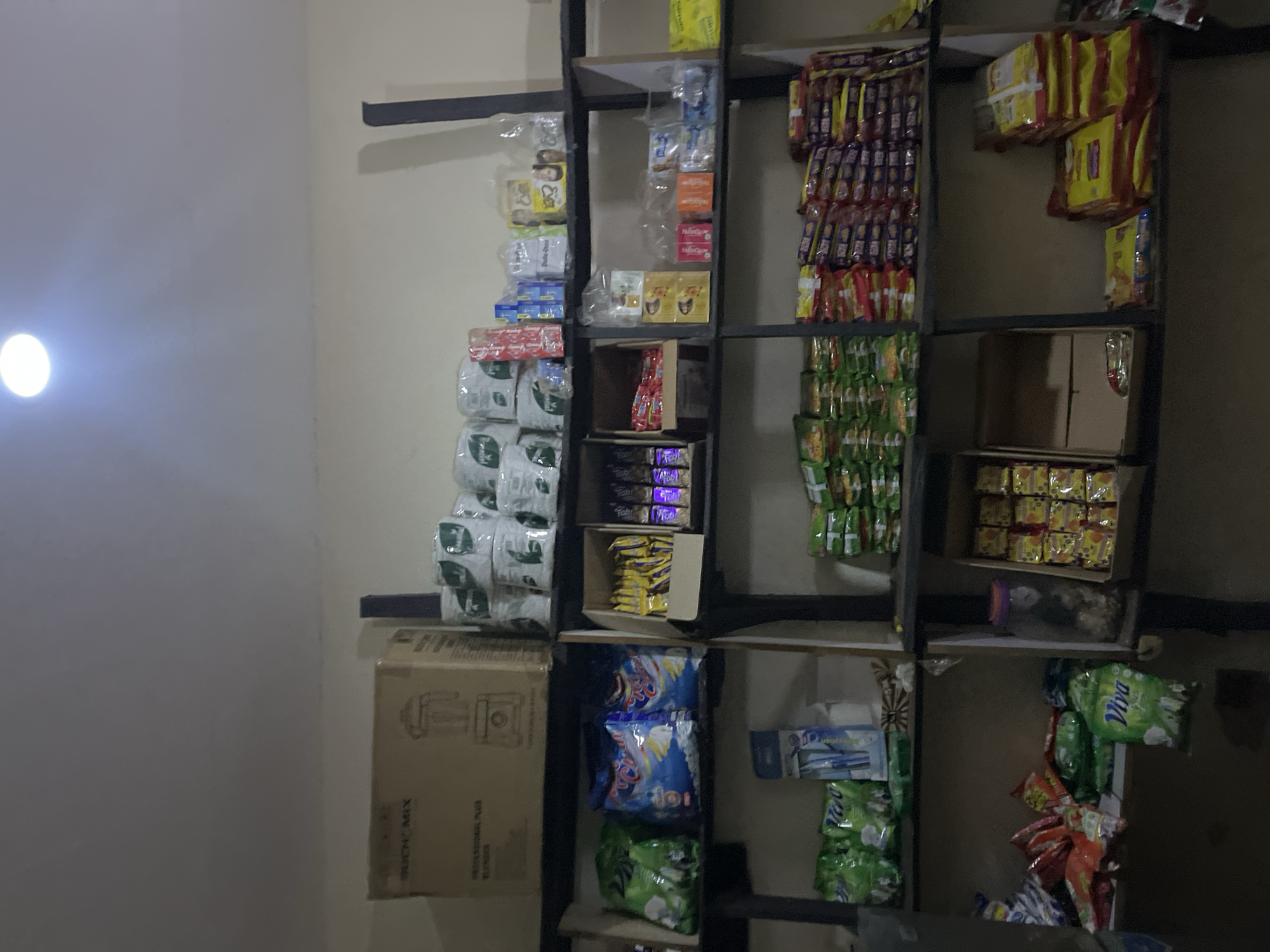 The image shows a convenience store in Abuja, Nigeria. The store has shelves stocked with various food items, snacks, and household goods. The shelves are made of metal and are organized with different items on each shelf. Items include packets of cookies, chips, beverages, and household supplies. The store appears to be in a well-lit area, although there is a glare from a light source.