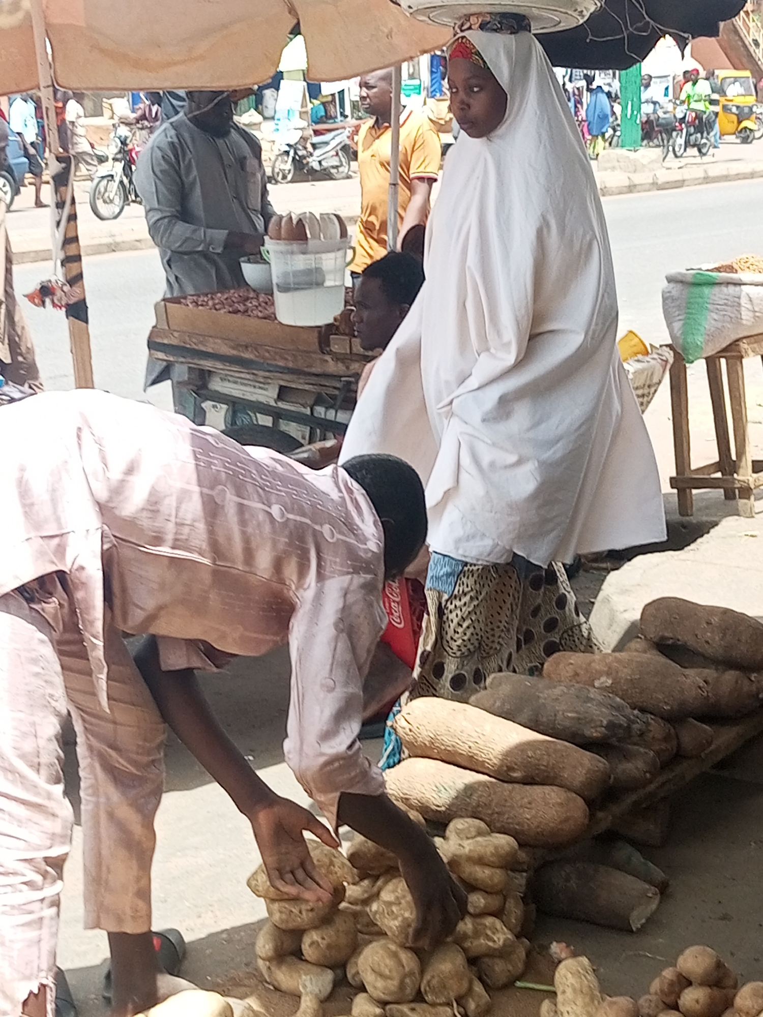 The scene is an open-air market in Nigeria. A woman wearing a white hijab stands behind a display of yams. A man in a pink shirt is picking through a pile of smaller tubers. Behind them are other vendors with peanuts and coconuts, as well as a road with motorcycles and a tricycle. The location is the city of Tempure.