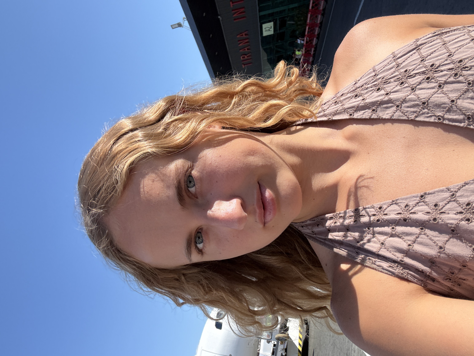 A young woman with blonde, wavy hair takes a selfie outdoors. She is wearing a light brown, patterned top. The background shows a bright blue sky and a building with the text "TIRANA INT" visible, suggesting an airport or similar transportation hub. The woman is looking towards the camera with a neutral expression. Sunlight is casting shadows on her face and clothing, indicating it is daytime. There's a part of an airplane visible at the bottom of the image. The overall impression is a casual, sunny day outing.