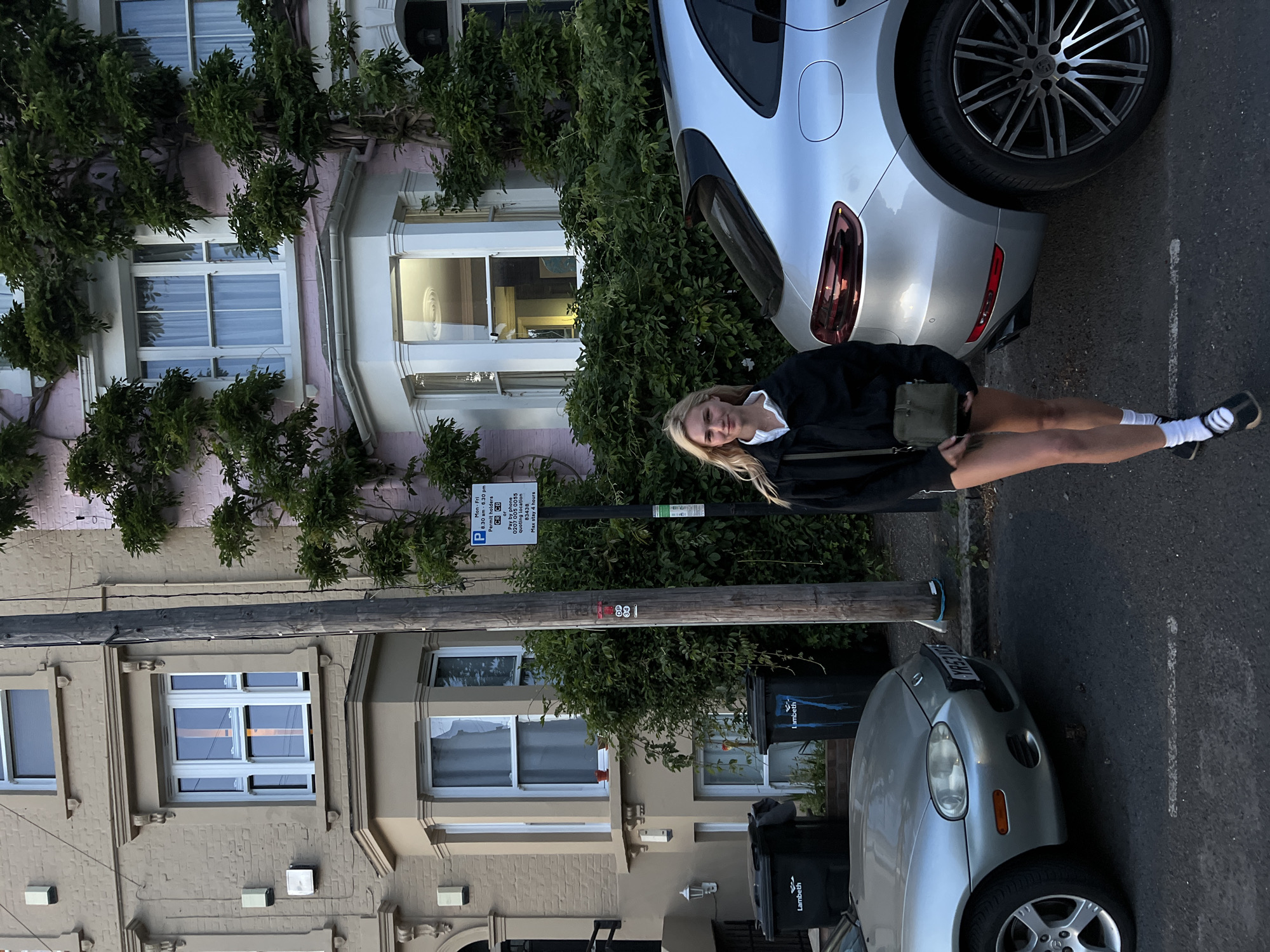The scene depicts a street in London, United Kingdom. A young blonde woman stands on the sidewalk next to a parked silver car and a partially visible silver Porsche. She wears a black jacket, shorts, white socks, and black shoes. She also has a green crossbody bag. A signpost is behind her. The houses lining the street are made of stone and brick, with windows and greenery along the walls. Two black trash containers are next to the street. The street has parking signs on it. The image is oriented sideways.