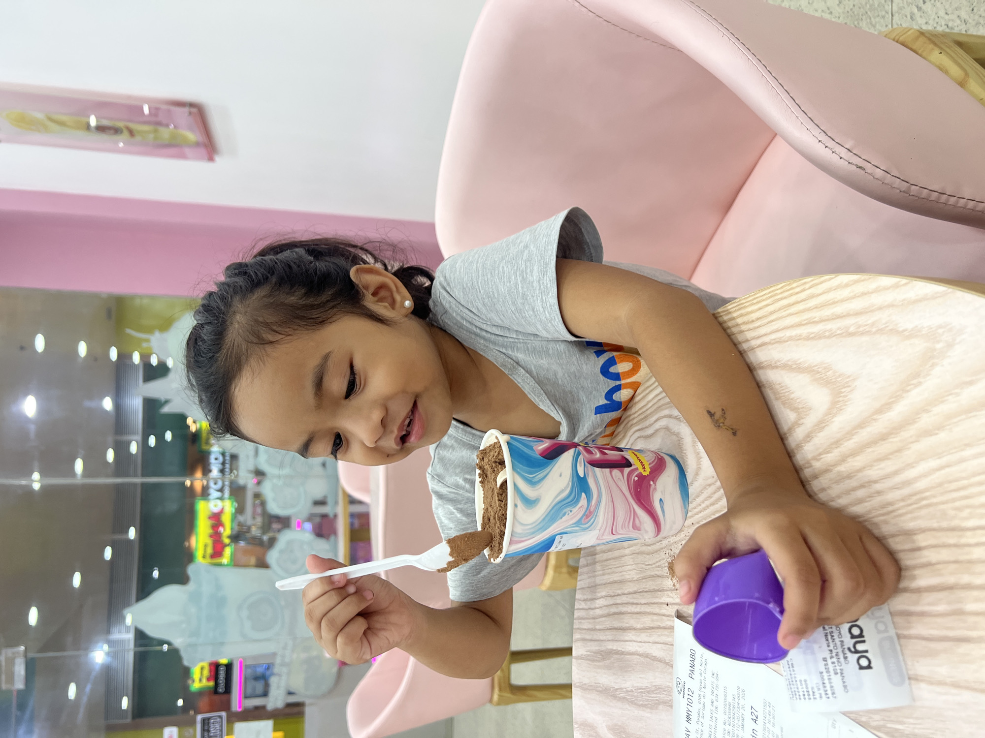 A young girl with dark hair pulled back in a ponytail sits at a wooden table in an ice cream parlor. She is wearing a gray t-shirt and holds a white plastic spoon in her right hand, which is scooping chocolate ice cream from a colorful cup. Her left hand is holding a small purple lid. On the table in front of her is a receipt. The girl has a small temporary tattoo of a dragon on her left forearm. The background shows pink chairs and a glass wall with cartoon ice cream cones and the word "GYCMON" visible through it. The location is identified as Panabo City, Philippines, specifically on the receipt which mentions "SAVIA ST, PANABO, 8105 BAYOG DEL NORTE".