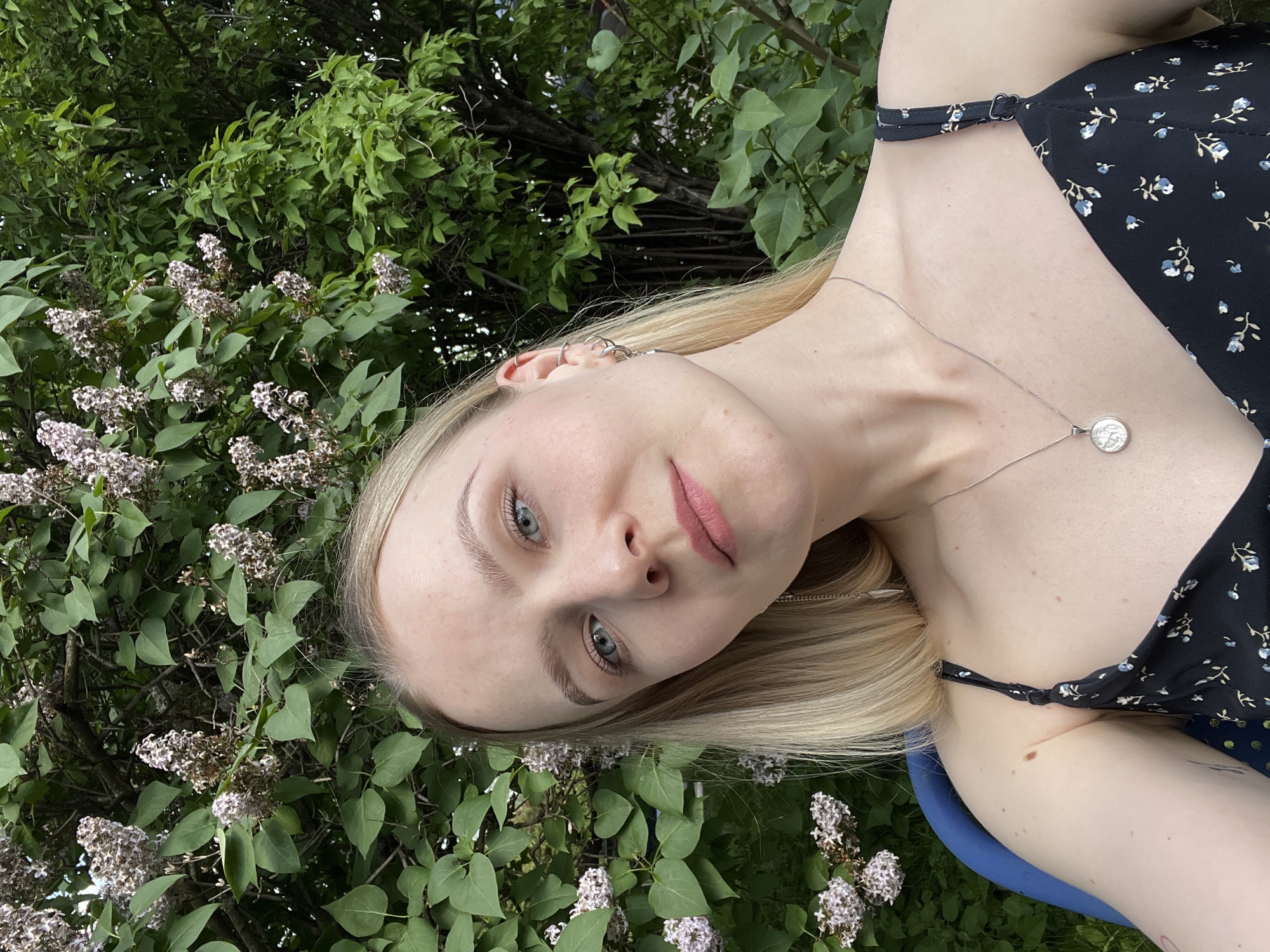 A young woman with blonde hair and fair skin is depicted in an outdoor setting. She is wearing a black dress with a floral pattern and a silver necklace. Her facial features include light eyes, pink lips, and a neutral expression. She has multiple earrings on her left ear. Behind her are flowering bushes and green foliage. Based on the provided information, the location is claimed to be Khimki, Russia, but there is no verifiable information in the image to support this.