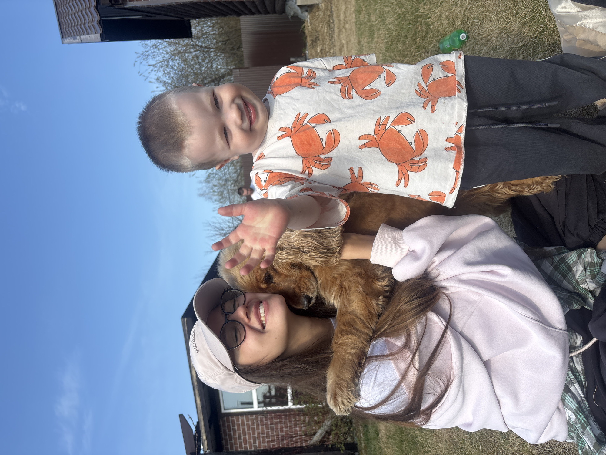 A young child and a young woman are in an outdoor setting, likely a backyard or park, with a dog. The child is a boy with light hair and a smiling expression. He is wearing a white shirt with orange crab designs and black pants. His right hand is raised, as if waving. The young woman is wearing glasses and a light-colored baseball cap. She has long brown hair and is wearing a light pink hooded sweatshirt. She is smiling and appears to be holding the dog. The dog is a Cocker Spaniel with long, wavy brown fur. The woman appears to be hugging the dog. The background shows a blue sky and a mix of grassy areas and some structures, including a building with a brick facade and a wooden structure. There is also a green bottle on the ground. The location is Emel'yanovo, Russia.