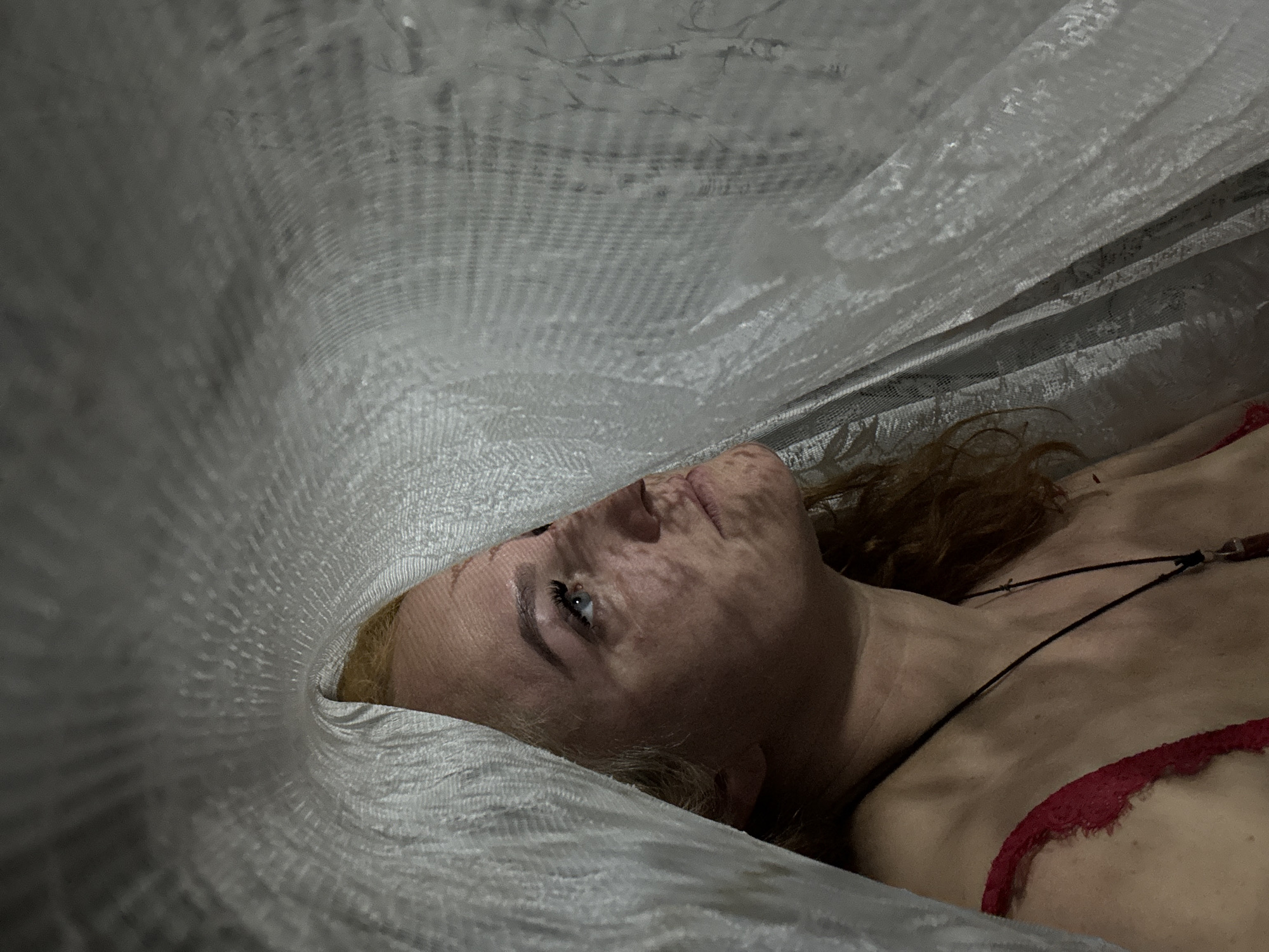 A pale-skinned woman with auburn hair lies supine under a white, textured material. She wears a red lace garment. A necklace is visible on her chest. Her head is turned to the left, and her eye is open. Shadows pattern her face. The location and other specifics cannot be determined from the image.