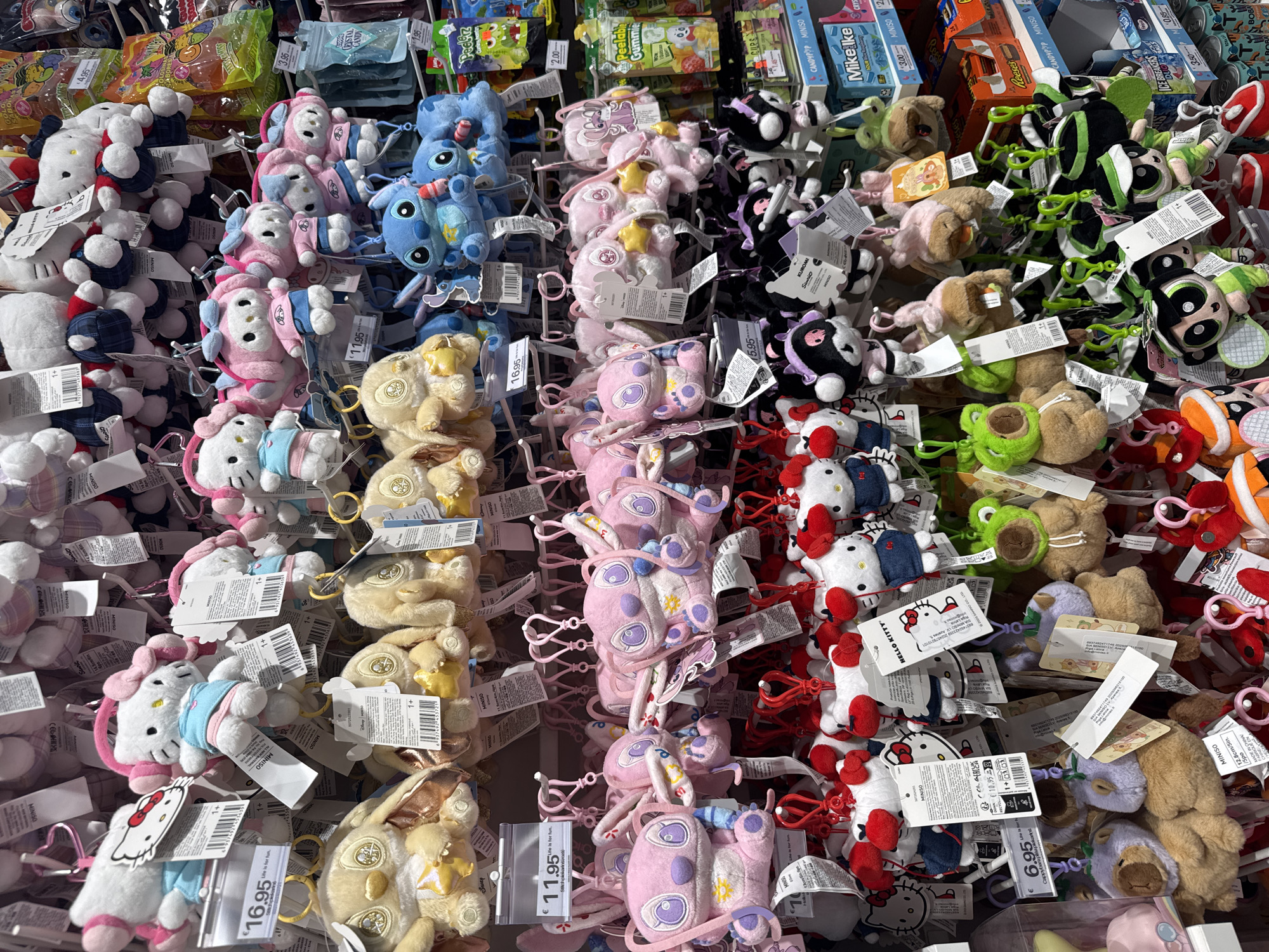 This media file captures a brightly lit retail display showcasing numerous small plush keychains and candies, likely within a store in Riga, Latvia. The scene is dominated by rows of plush characters hanging on white metal racks.
The primary subjects are licensed plush keychains from popular franchises. On the left, numerous white Hello Kitty plushies are visible, many adorned with red bows and some wearing blue or pink outfits. Moving right, there are pink plushies resembling My Melody or Angel, followed by blue Stitch plushies. Further to the right, black and white Kuromi plushies with purple accents are seen. Additional plushies include green frogs and brown characters, identified by a visible tag as "Cappybara" (capybara).
In the background, above the plushies, various packaged candies and snacks are displayed. Visible brands include "Mike Ike," "Peelabl Gummie," "CRYSTAL CANDY," "Reese's CUPS," "ICE BREAKERS DUO," and "BOBA" cans, suggesting a general store or a section dedicated to novelty items and treats.
Notable details include numerous price tags indicating costs in Euros, such as "€16.95" for Hello Kitty and Kuromi, "€11.95" for Stitch and Angel, and "€6.95" for the capybara plushies. Candy prices range from "€2.00" to "€4.95." Many tags prominently feature "MINISO," indicating the retailer or brand. Other visible text includes "MADE IN CHINA," "FABRIQUE EN CHINE," age ratings like "1+," and various product numbers and barcodes. The items are tightly packed on the display, suggesting a full stock. No people or specific activities other than the merchandise display are visible. The artificial lighting indicates an indoor setting.