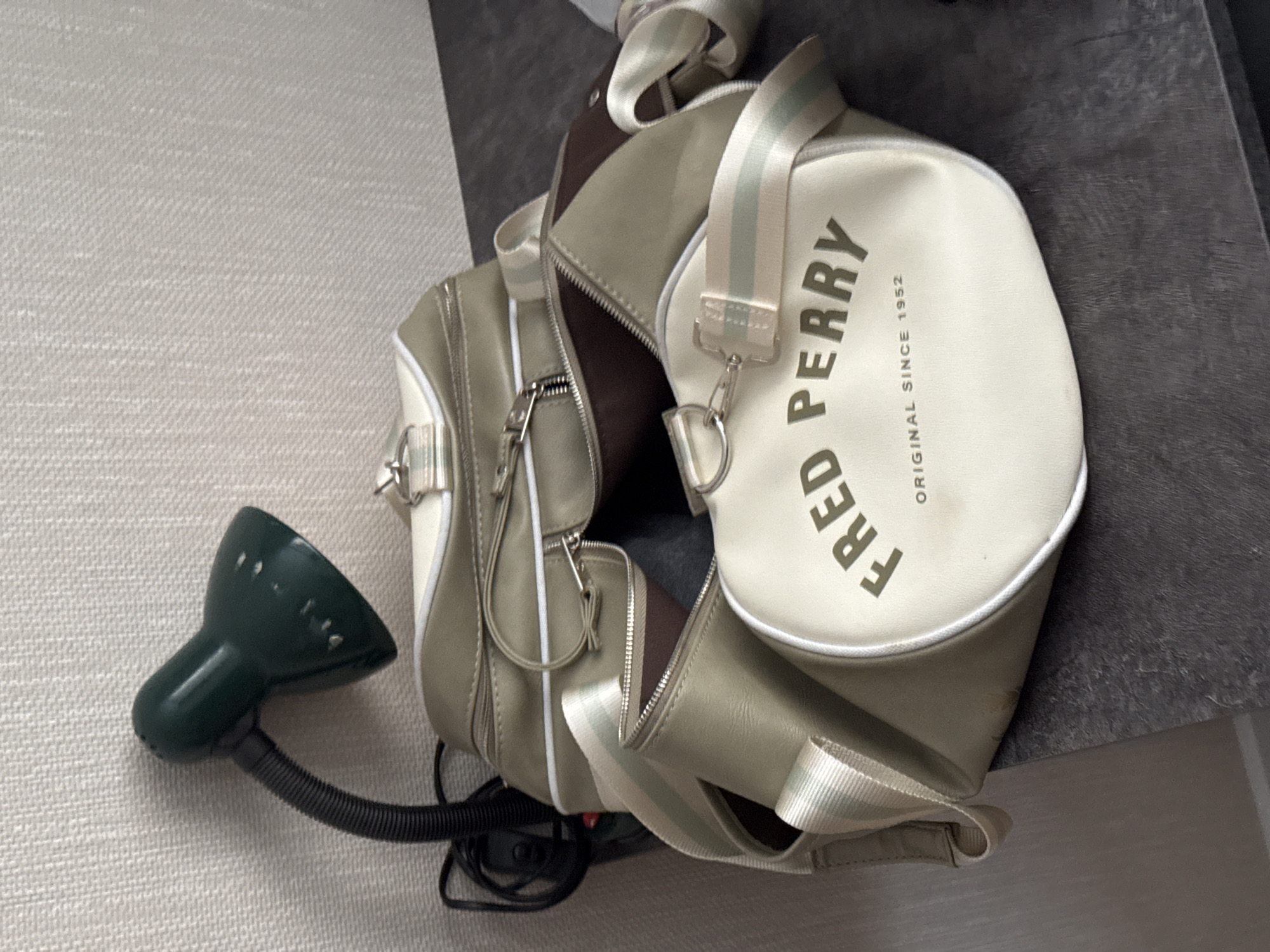 The image shows a close-up of a light green and cream duffel bag with the "FRED PERRY ORIGINAL SINCE 1952" logo on its front. The bag has various zippers and straps, some of which are a cream color with light green and white stripes. Next to the bag is a small, dark green desk lamp with a flexible neck and a black cord that trails off to the left. The background consists of a textured, light-colored wall and a dark, textured surface, likely a tabletop or shelf. The overall impression is of a domestic or retail setting. There is no visible indication of the city of Perm, Russia.