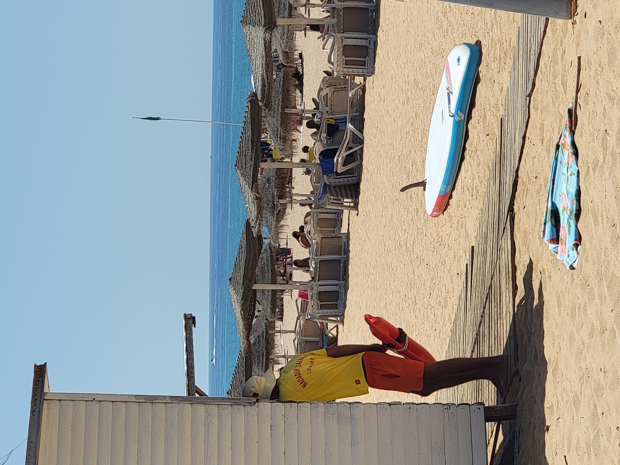 The image depicts a sunny day at Quarteira beach in Portugal. In the foreground, a lifeguard wearing a yellow t-shirt with "NADADOR SALVADOR LIFEGUARD" printed on it and orange shorts stands by a white wooden beach hut. He is holding a red rescue buoy. A stand-up paddleboard with blue and white colors lies on the sandy beach, partially on a wooden walkway. Further back, beach umbrellas and lounge chairs are set up, with several people relaxing under them. The ocean stretches to the horizon under a clear blue sky, with a small boat visible in the distance. A dark flag on a pole stands near the water's edge. A folded beach umbrella with a floral pattern rests on the sand in the lower part of the image.