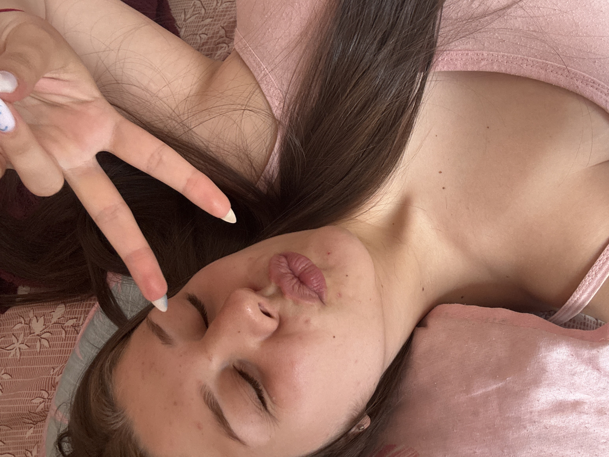 The media file is a close-up, top-down, selfie-style shot of a person lying down. The subject is a young person, likely female, with long, dark brown hair. They are wearing a pink camisole or tank top. Their eyes are closed, and their lips are pursed, resembling a kiss. One hand is raised, with the index and middle fingers extended in a peace sign, and the thumb is tucked in. The nails are painted white and have a light blue design. The background appears to be a bed or couch with a patterned fabric in muted pink and beige tones. There is a glimpse of a pink pillow on the right side of the frame. The lighting suggests it is daytime, possibly indoors. The overall mood is relaxed and playful. There is no visible text in the image.