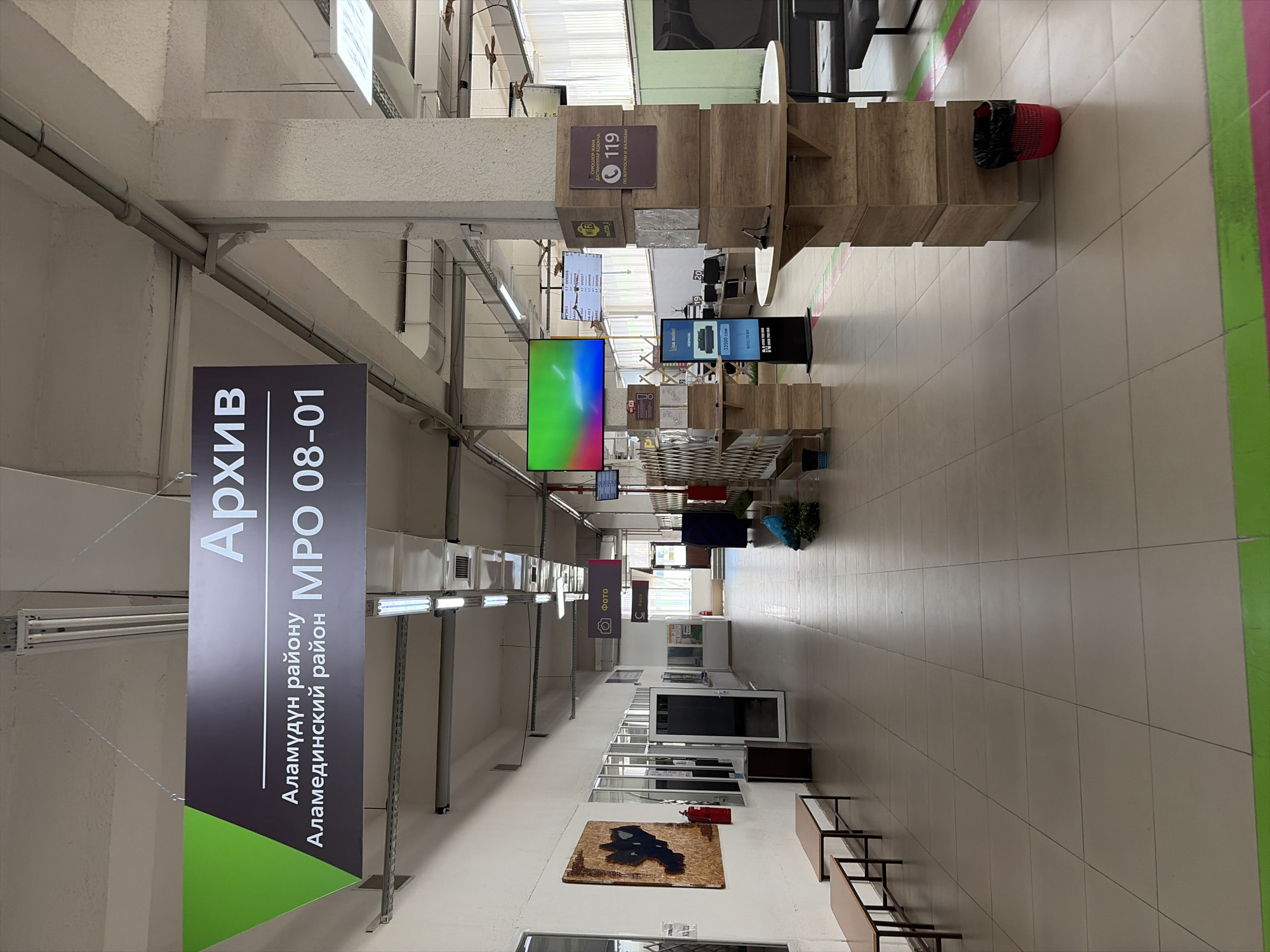 The image depicts a wide, indoor hallway with tiled flooring. A large sign overhead reads "Архив" (Archive) in large white letters, with smaller text below indicating "Аламудун району" (Alamudun district) and "Аламединский район" (Alamediynsky district) next to "MPO 08-01". Along the left wall, there are several wooden benches and a large piece of art. Further down the hallway, there are glass-fronted offices, a fire extinguisher, and various signs, including one with a camera icon and the word "ФОТО" (Photo), and another labeled "Касса" (Cashier). On the right side of the hallway, there is a prominent pillar covered with wooden panels, featuring a "Wi-Fi" logo, a number "119", and text in what appears to be Kyrgyz. There are also digital displays, furniture, and trash receptacles visible throughout the scene. The overall environment suggests a public building, possibly an administrative office or a service center.