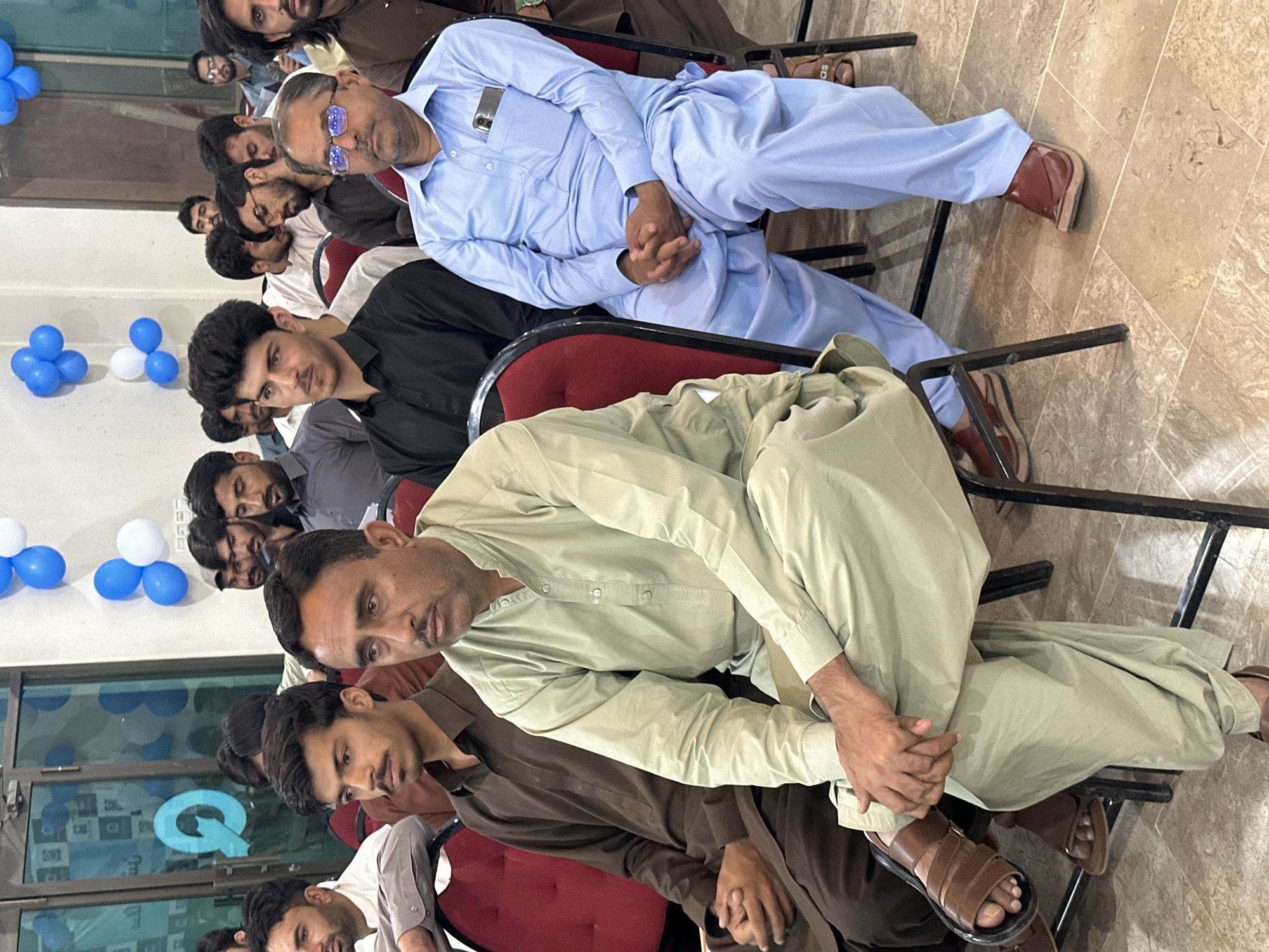 A medium shot captures a group of men sitting in chairs in what appears to be an indoor gathering space. The men are dressed in traditional South Asian clothing, mostly shalwar kameez, in various neutral and light colors. Their postures suggest they are attending an event or meeting. Some men are looking forward, while others have their gaze directed elsewhere. The environment is a well-lit room with patterned floor tiles and balloons visible in the background, suggesting a festive or celebratory occasion. The image is likely taken in Quetta City Tehsil, Pakistan, given the context of the location.