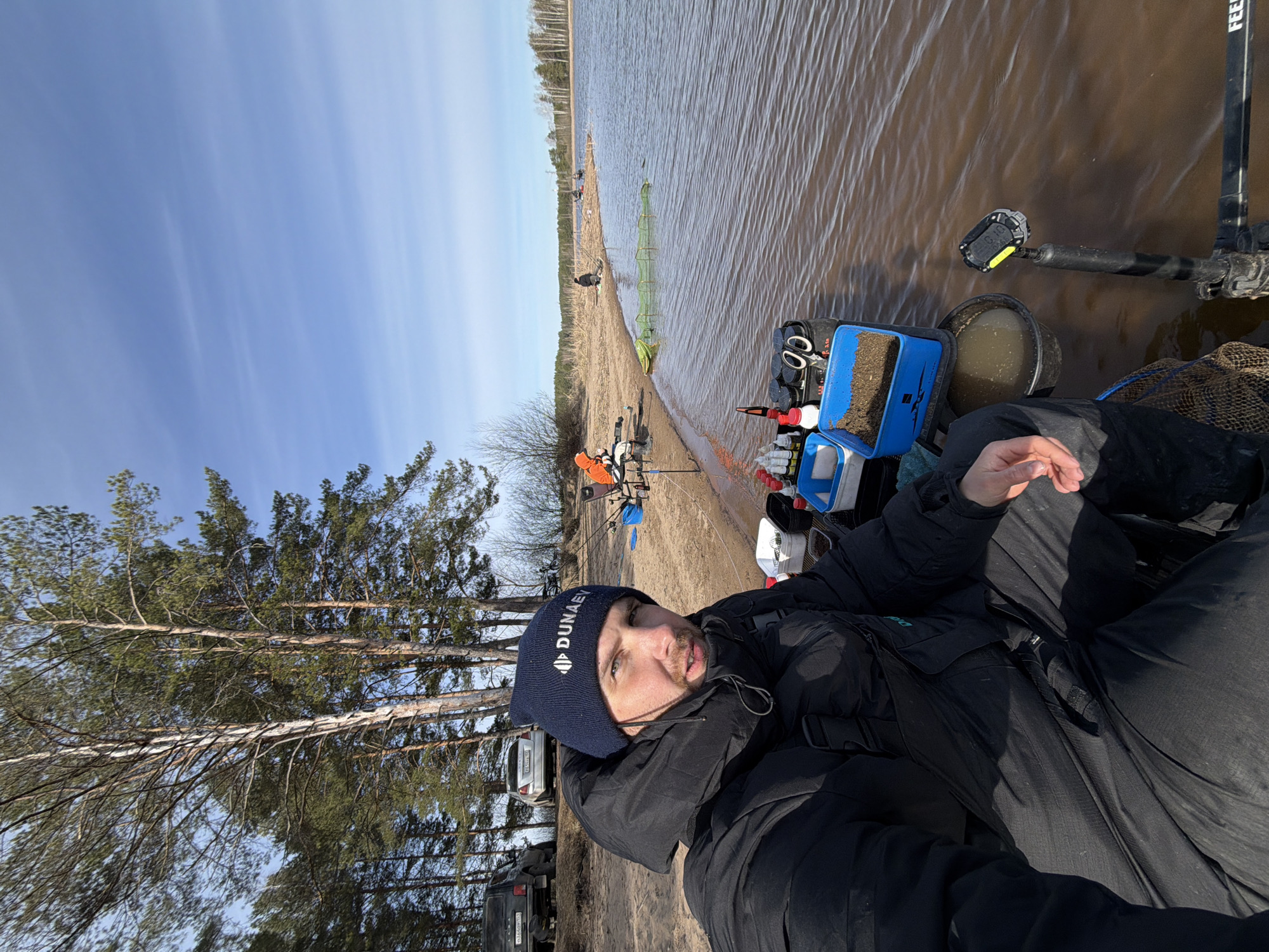 This media file presents an outdoor fishing scene from a first-person perspective on a clear day in Chaikovsky, Russia. The main subject, a man with a beard, takes a selfie, looking directly at the camera with a neutral expression. He wears a black puffy jacket and a dark blue beanie with the word "DUNAEV" and a diamond logo.
He is positioned on a sandy or dirt bank next to a calm body of water, likely a large river or lake. To his right, various fishing equipment is laid out, including blue containers filled with bait or groundbait, a black bucket, and a black fishing rod stand with "FEEDE" (likely "FEEDER") visible. An electronic device, possibly a Garmin fish finder or transducer, is mounted on a stand in the water, showing "GARMEN" and some numbers. Fishing rods are visible set up along the bank.
In the mid-ground, other individuals are scattered along the shore, also engaged in fishing activities, with some standing by the water's edge and what appears to be a long net or fish-keeping sack partially submerged. The far bank is lined with a sparse forest of coniferous trees. The sky is bright and clear blue, indicating sunny weather. In the background, nestled among the trees, two vehicles, including a black SUV, are parked. Part of a license plate on one vehicle shows "24" and "72". The overall atmosphere is peaceful, depicting a fishing trip on a sunny, possibly cool, day.