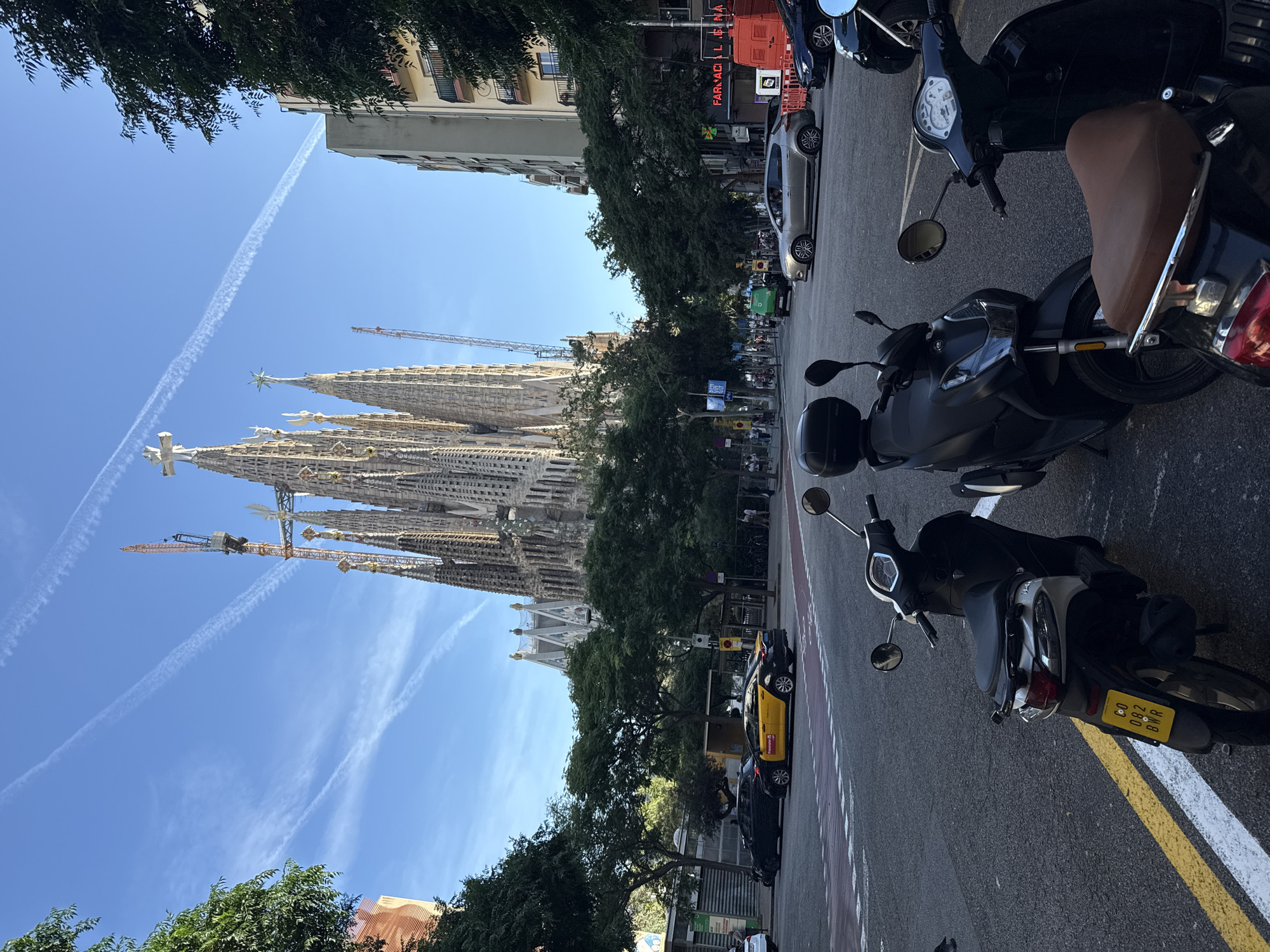 This image captures an urban street scene in Barcelona, Spain, dominated by the Sagrada Familia basilica in the background. In the foreground, three scooters are parked on the right side of a paved street. The leftmost scooter has a yellow license plate reading "G0 082 BWR". The street features double yellow lines, white dashed lines, and red-painted sections, indicative of a bike lane or special road marking.
The midground depicts an active street intersection. A yellow and black taxi, marked with "FREE NOW," is visible, along with a silver hatchback, a dark sedan, and a green delivery vehicle. Pedestrians can be seen on sidewalks lined with numerous mature green trees. On the right, a multi-story building displays a red "FARMACIA" sign, and a red-orange delivery truck is parked nearby. Another building with balconies is visible further down the street on the right.
The background is defined by the Sagrada Familia, characterized by its intricate, towering spires. Multiple large construction cranes are positioned around the basilica, signifying ongoing construction work. Surrounding the basilica are other multi-story city buildings. The sky is clear blue with several distinct white airplane contrails. The scene is illuminated by bright daylight, casting visible shadows.