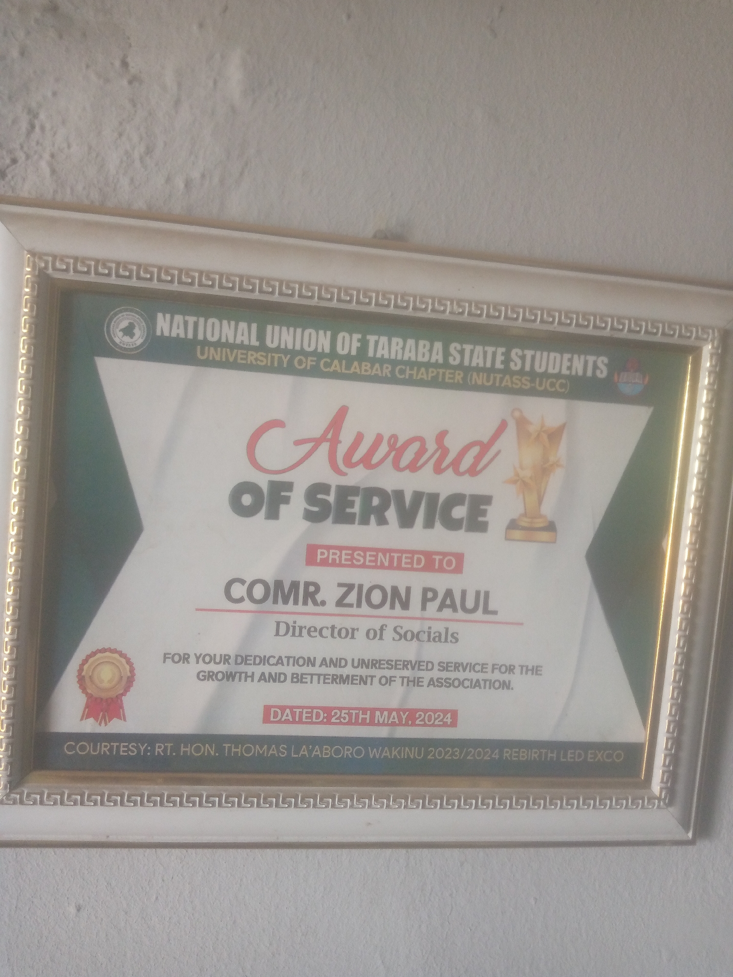A framed award for service is centrally displayed, hanging against a light-colored, textured wall in Jalingo, Nigeria. The frame is white with an intricate gold Greek key pattern along its inner edge. The award itself, printed on a white background with green diagonal sections in the upper corners, is titled "Award OF SERVICE" in red script and bold black capital letters, respectively. A gold trophy emblem is positioned in the upper right quadrant of the award, while a gold seal or medal emblem is in the lower left.
The award is "PRESENTED TO COMR. ZION PAUL" for his role as "Director of Socials," acknowledging "YOUR DEDICATION AND UNRESERVED SERVICE FOR THE GROWTH AND BETTERMENT OF THE ASSOCIATION." The awarding body, prominently displayed in a green banner at the top, is the "NATIONAL UNION OF TARABA STATE STUDENTS UNIVERSITY OF CALABAR CHAPTER (NUTASS-UCC)," flanked by two circular emblems. The award is "DATED: 25TH MAY, 2024" and states "COURTESY: RT. HON. THOMAS LA'ABORO WAKINU 2023/2024 REBIRTH LED EXCO" at the bottom. The wall behind the frame shows subtle imperfections and a rough, possibly stucco-like texture.