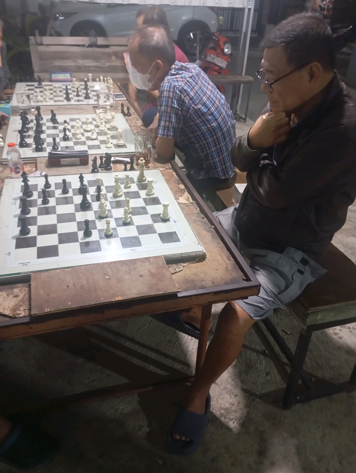 The image depicts an outdoor setting in Tangerang, Indonesia, likely at dusk or night, with artificial lighting illuminating the scene. Two men are engaged in playing chess at separate tables.
In the foreground, a man wearing glasses and a dark jacket sits on a wooden bench to the right of the frame. He is leaning forward, with his chin resting on his hand, appearing to be contemplating his next move. In front of him is a chessboard with black and white pieces. The table he is using is weathered wood, with a metal frame.
Further back, another man, partially obscured, is seated at a table, also playing chess. He is wearing a checkered shirt and a white face mask. His chessboard is also set up with pieces.
The environment appears to be a casual gathering place, possibly a street-side stall or an open-air recreational area. In the background, indistinct shapes suggest vehicles, including what might be a red motorcycle. The ground is paved, and shadows are cast across it, indicating the lighting sources. The overall impression is one of a relaxed, perhaps competitive, game of chess in a local Indonesian setting.