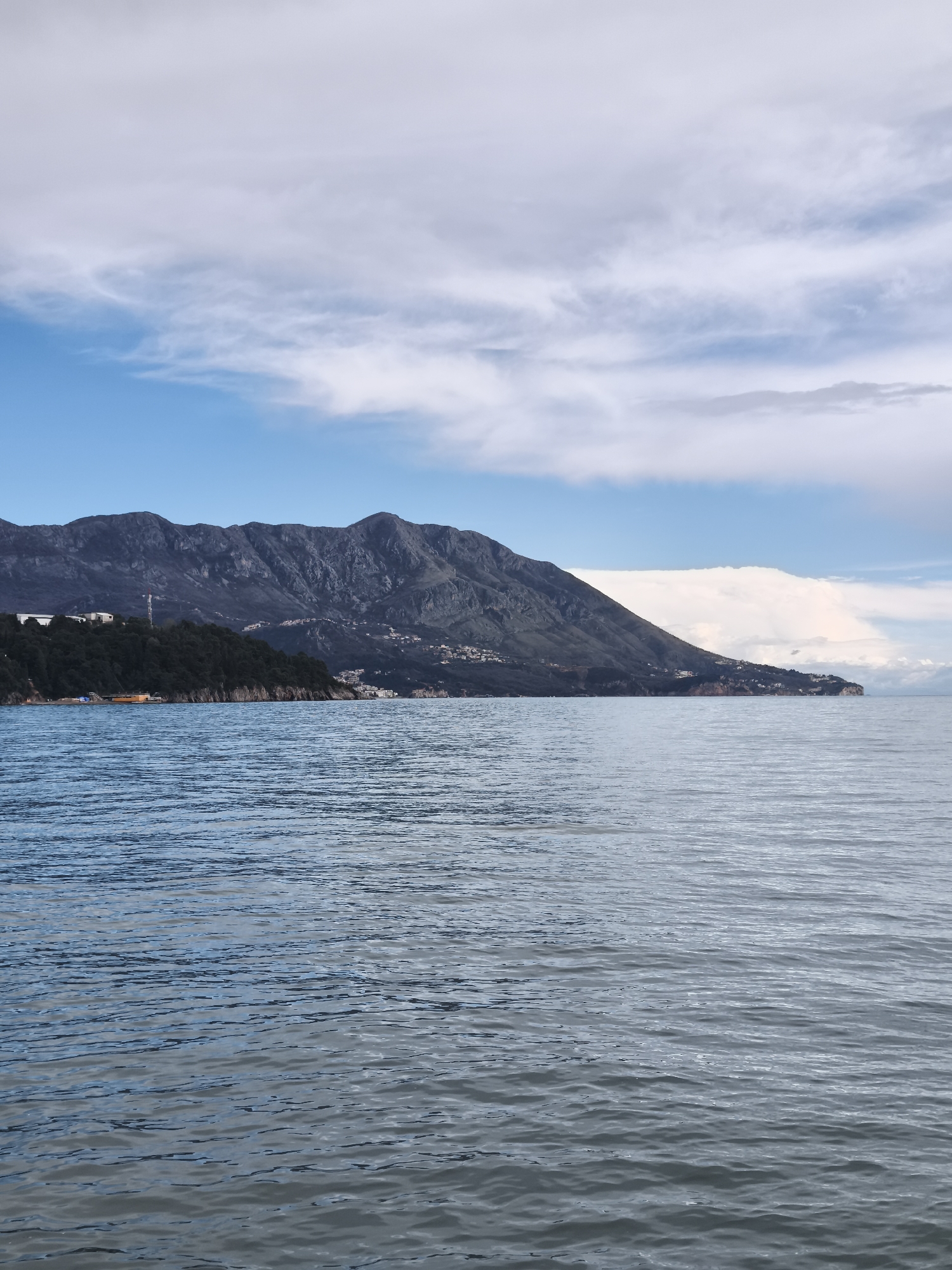 The image depicts a panoramic view of the Adriatic Sea and coastline in Budva, Montenegro. The scene is dominated by a vast body of water, characterized by ripples, occupying the lower half of the frame, exhibiting shades of blue-gray. No vessels or individuals are present on the water.
The middle ground features a rugged, dark mountainous terrain extending horizontally across the frame. On the left side, a prominent, densely forested headland protrudes, partially obscuring the mountain base. Visible on this headland are a white building complex near the top of the treeline and a tall, red and white striped telecommunications mast. Closer to the water's edge on the left, a long, yellow-orange building with a dark roof is situated adjacent to a small, light-colored beach area.
Numerous white and light-colored buildings are scattered along the lower slopes of the mountains, indicating human habitation. The skyline is composed of a mixture of clear blue sky and diverse cloud formations, including thin, wispy cirrus clouds and larger, denser stratocumulus clouds. A substantial, bright white cloud mass is present above the sea on the far right. The overall lighting suggests daytime conditions. No people, explicit actions, or interactions are visible in the scene.