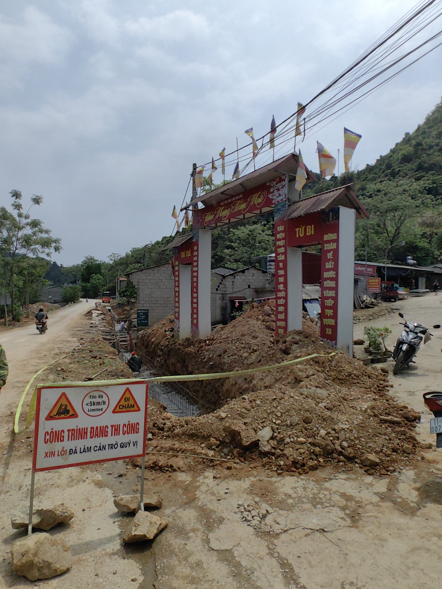 The image shows a construction site on a road in Xã Đàm Thủy, Vietnam. A large trench has been dug across the road, with a pile of dirt beside it. A sign in Vietnamese warns of the construction and a speed limit of 5 km/h. In the background, a decorative gate with religious flags stands near some buildings. People are working in the trench, and motorcycles are parked nearby and driving on the road. The environment includes trees, a mountain, and power lines overhead.