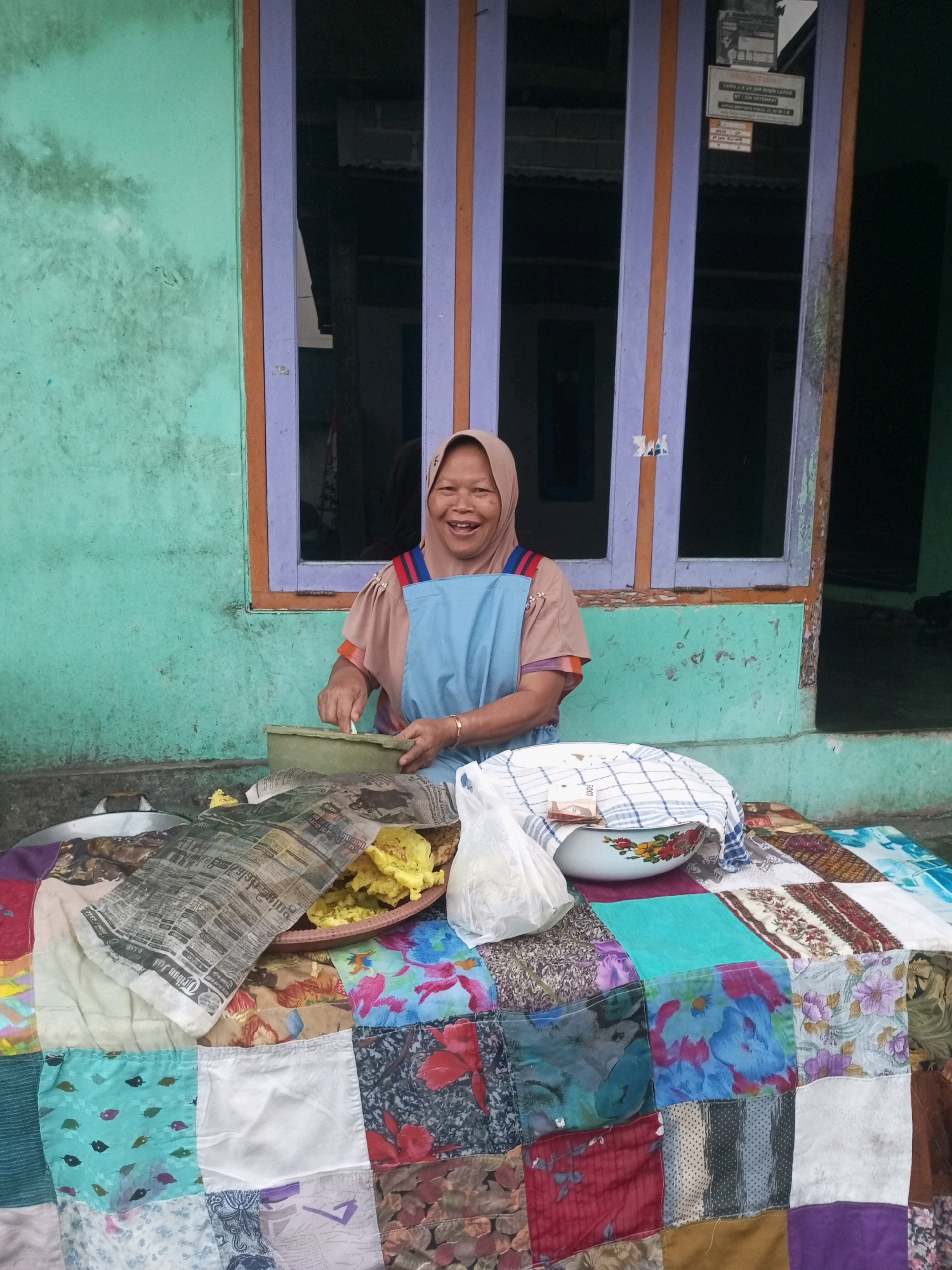 In Andongsili, Indonesia, a woman wearing a light brown headscarf and a blue apron smiles at the camera while preparing food. She is seated in front of a turquoise-colored wall with a window featuring purple and orange-painted vertical bars. In front of her is a table covered with a colorful patchwork cloth. On the table, a green bowl, a basket of fried food wrapped in newspaper, and a white plastic bag are visible. A ceramic bowl with a floral pattern, covered with a checkered cloth and containing a bill, is also present. The scene suggests a street food vendor setting up for business.