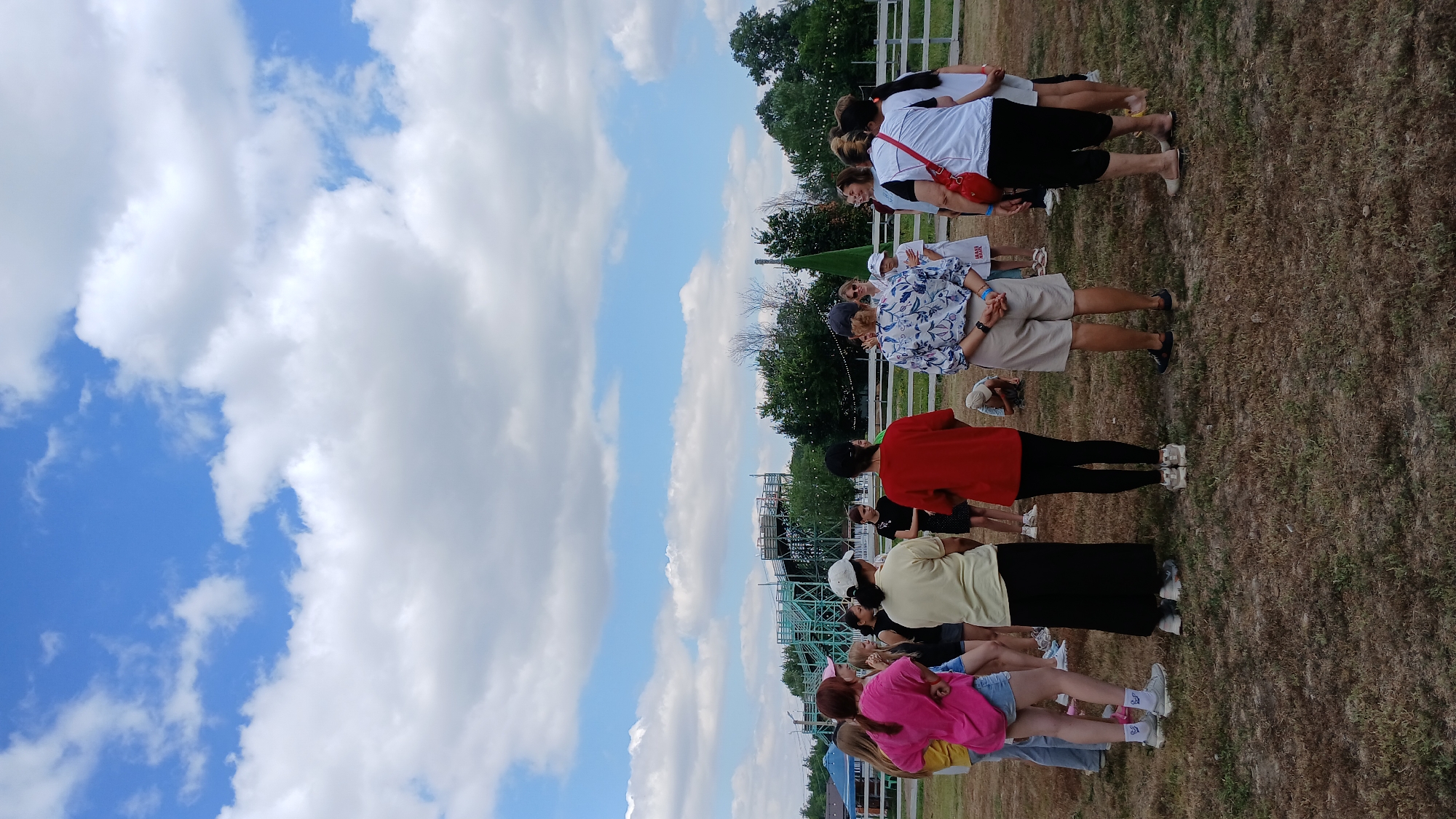 In Derköl, Kazakhstan, a group of approximately twelve people stand in a circle on a grassy field under a partly cloudy sky. The individuals, appearing to be a mix of adults and teenagers, are facing inwards and engaged in conversation or instruction. Their attire varies, with some wearing casual clothing like t-shirts, shorts, and jeans, while others are dressed in more formal wear. The environment suggests an outdoor event or gathering, possibly at a park or recreational area, with a structure resembling a roller coaster or amusement park ride visible in the background, along with trees and fencing. The ground is covered in dry grass, indicating a warm climate.