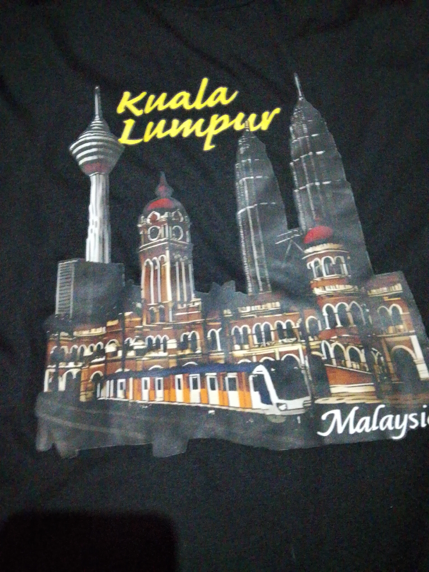 The image shows a black t-shirt with a print depicting the Kuala Lumpur skyline. Prominent landmarks include the KL Tower, the Sultan Abdul Samad Building, and the Petronas Twin Towers. A train is depicted in front of the building. The words "Kuala Lumpur" are printed in yellow above the skyline and "Malaysia" is printed in white below. The scene is a representation of Kuala Lumpur, Malaysia, not Bukit Ketok, Indonesia.