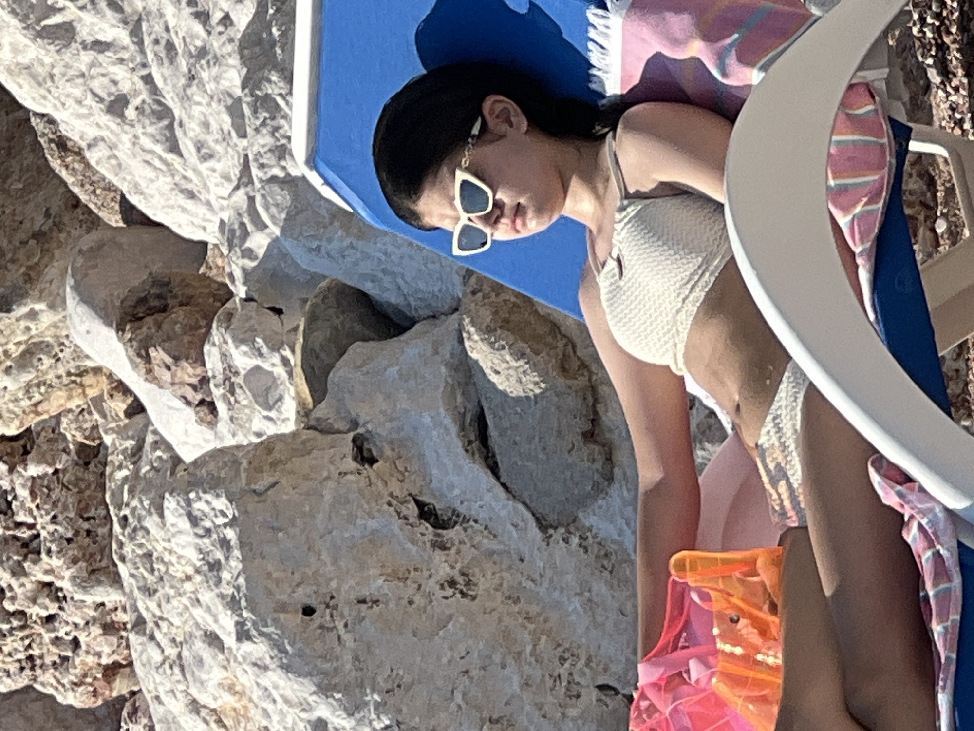 A person is lying on a beach chair under a blue umbrella. They are wearing sunglasses and a white bikini. The background is rocky and appears to be a beach. The sun is shining, suggesting it is daytime. A pink and white striped towel is draped over the back of the beach chair. There is an orange plastic bag in the foreground.