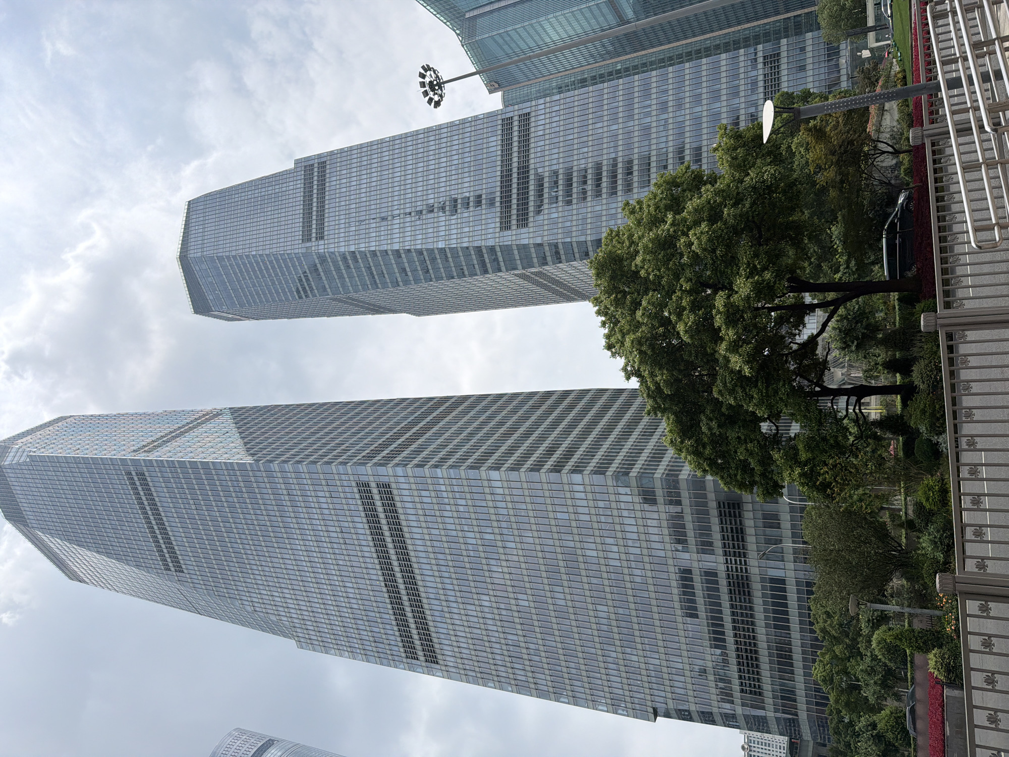In the city of Pudong, China, the image captures a wide-angle view of modern skyscrapers under a cloudy sky. Two prominent glass-fronted towers dominate the scene, with their sharp angles and reflective surfaces creating a sense of immense scale. The skyscraper on the left is taller and more slender, while the one on the right is slightly shorter and wider with a more octagonal top. In the foreground, a lush green tree with full foliage stands prominently, partially obscuring the lower sections of the buildings. Behind the tree, there is dense greenery, including manicured bushes and flower beds with red and yellow blossoms. A modern streetlamp with multiple circular lights is visible to the right of the tree, casting a soft glow. A light-colored metal fence with decorative patterns runs along the bottom of the frame, separating the foreground from a paved area and a street where a dark-colored van is parked. The overall impression is of a contemporary urban landscape with a blend of imposing architecture and curated natural elements.