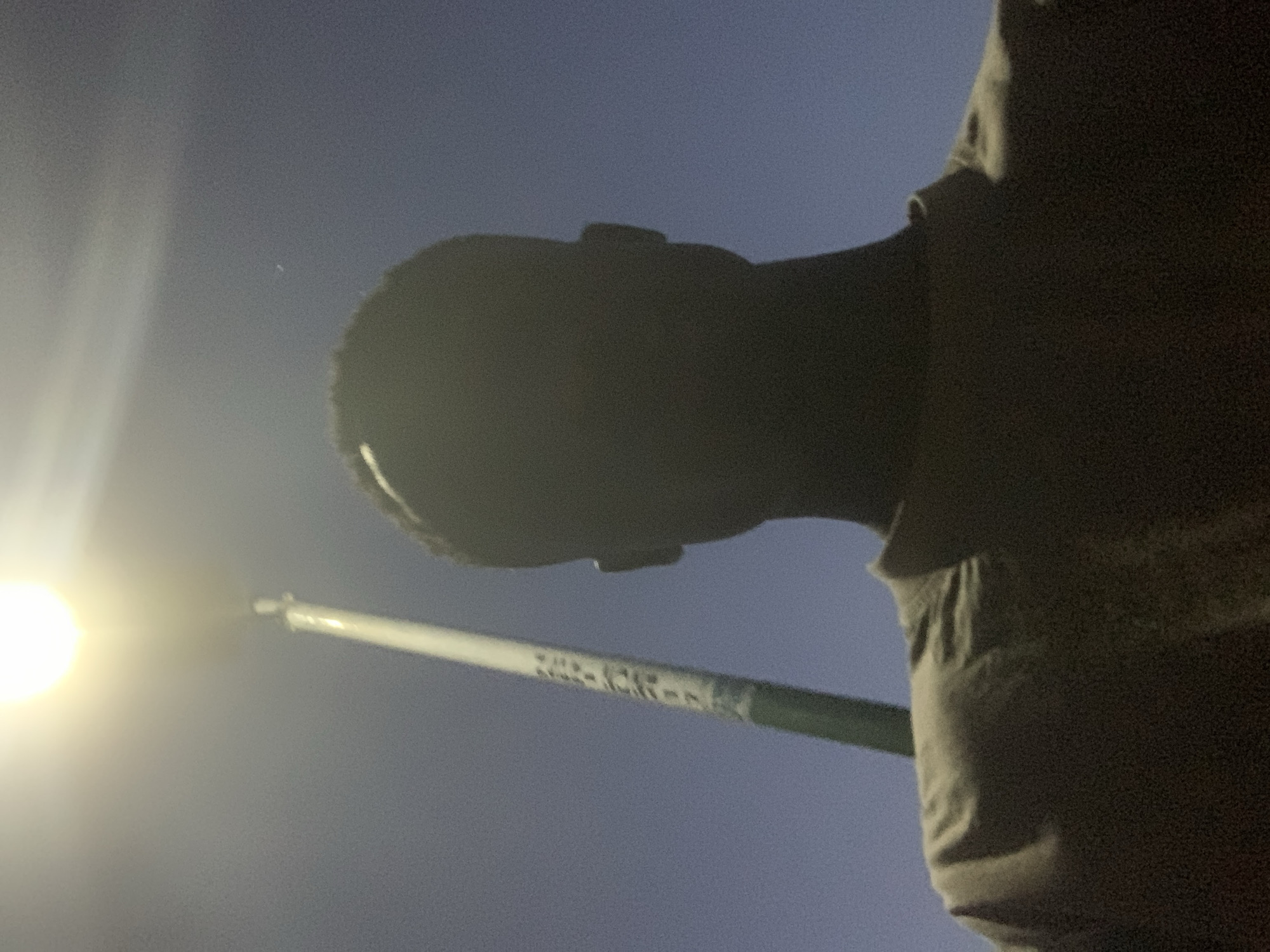 A low-angle shot shows a person's head and shoulder against a dark blue or grey sky. A light source, possibly a streetlamp, is visible to the left. A white pole with green accents, potentially a traffic light or other type of signpost, extends diagonally across the image, obscuring some of the sky. The person is wearing a dark-colored shirt or garment. The background is blurred and dark, suggesting a night scene. The location, Gbeleko, Nigeria, is unidentifiable from the image alone.