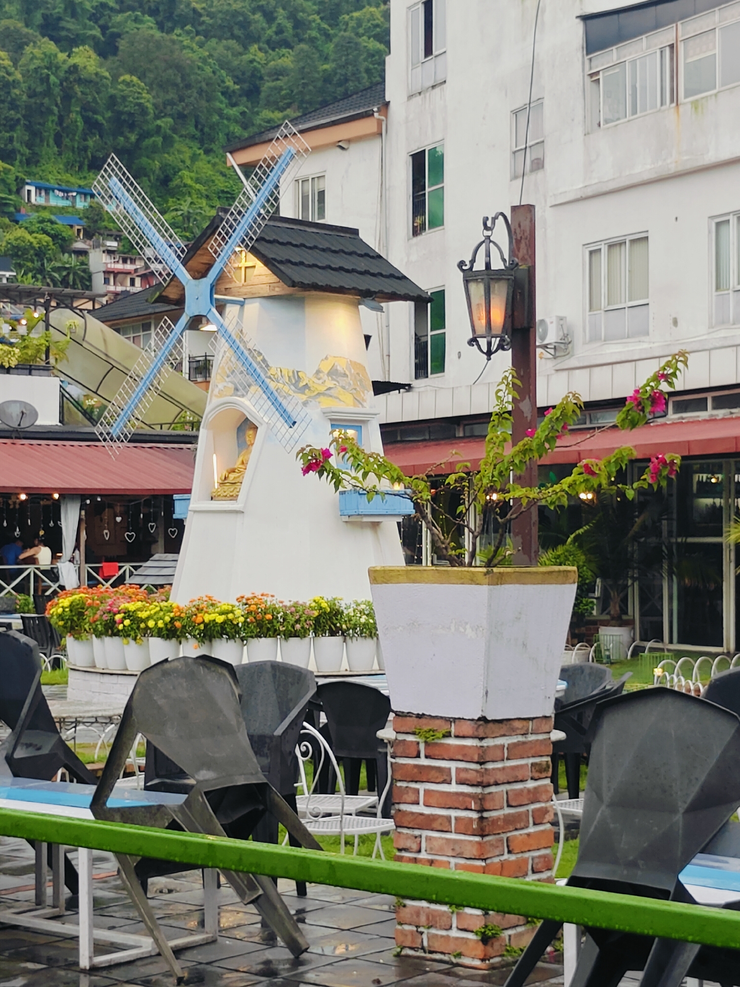 The image depicts an outdoor seating area, likely a restaurant or cafe, with a decorative windmill as a central feature. In the background, a multi-story building with white walls and numerous windows is visible, situated against a backdrop of lush green hills. The foreground shows several empty black chairs and tables, some of which appear to be made of metal. A planter with a potted plant with pink flowers stands prominently in the middle ground, mounted on a brick pillar. White flower pots filled with colorful flowers are arranged along the base of the windmill. The overall atmosphere suggests a serene, perhaps slightly overcast, day in a scenic location. This scene is consistent with the ambiance found in popular tourist areas of Pokhara, Nepal, known for its lakeside charm and mountainous scenery.