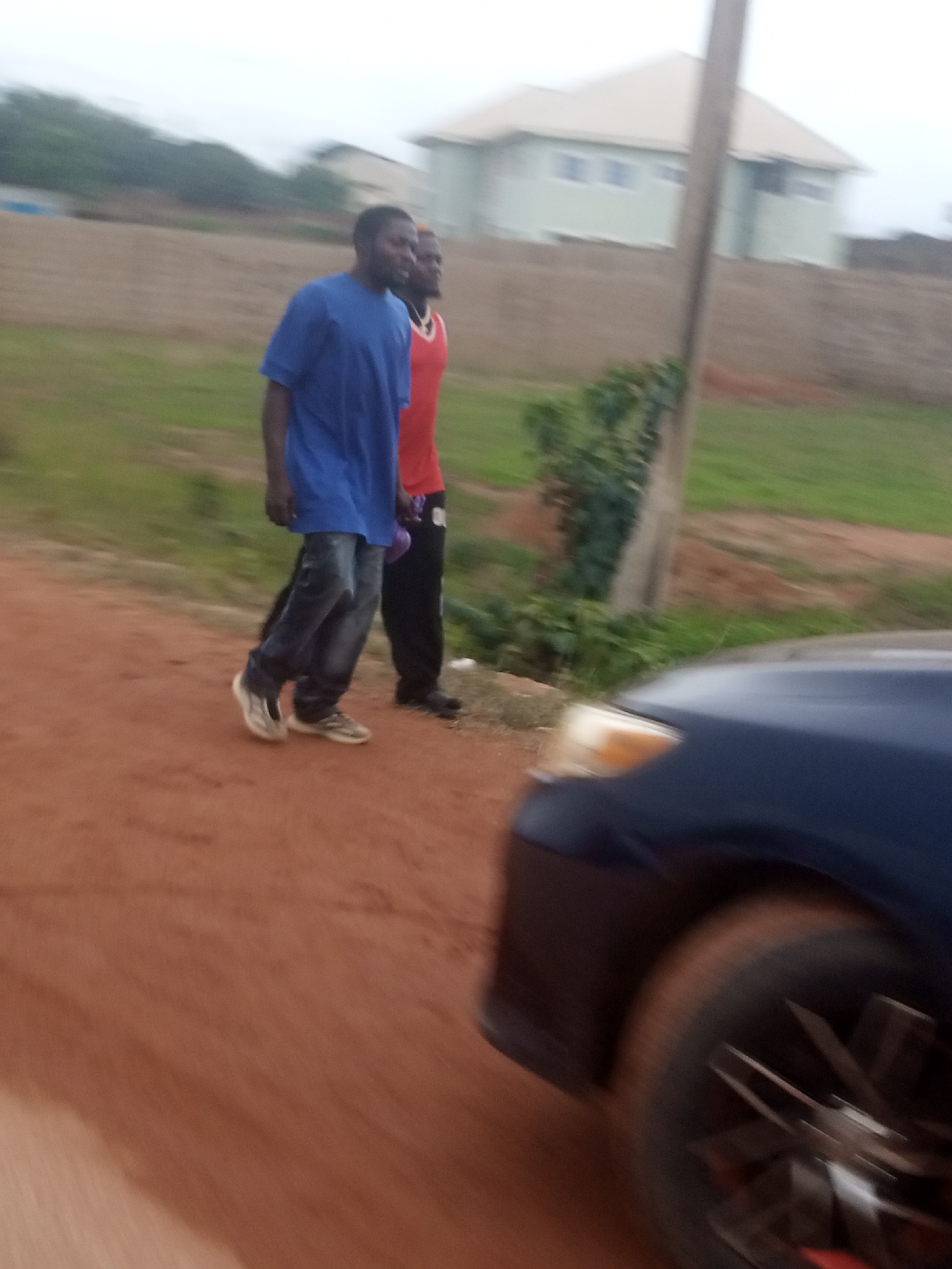Two men are walking on a dirt road. The man in the foreground is wearing a blue t-shirt and dark jeans, and is walking with his body angled towards the left. He is wearing white sneakers. Behind him, another man is walking, wearing a red sleeveless shirt and dark pants. He is walking slightly behind and to the right of the first man. The scene appears to be in a rural or semi-rural area. In the background, there is a light green building with a light-colored roof, possibly a house, and a fence. There is also green vegetation and dry, brown earth. A dark blue car is visible in the foreground, with its front bumper, headlight, and wheel in view. The image is slightly blurred, suggesting movement, possibly taken from a moving vehicle. The sky appears overcast, indicating a cloudy or possibly hazy day. The time of day is not clearly discernible due to the lighting conditions. The setting seems to be Kwang, Nigeria. No text is visible in the image.