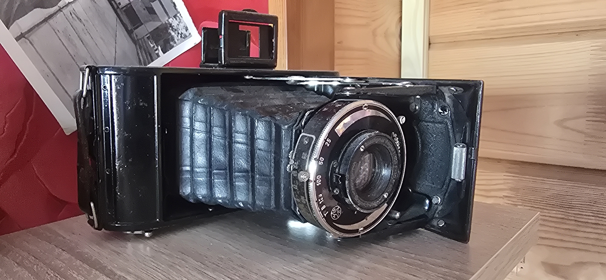 A black, vintage bellows camera is displayed on a light brown wooden surface. The camera is angled to the right, with its bellows partially extended and the lens assembly prominent. The lens has a circular dial with numerical markings and some text. A small black viewfinder is attached to the top of the camera. To the left of the camera, a portion of a black and white photograph is visible, leaning against a red textured surface. In the background, a light-colored wooden wall with horizontal planks is visible. The scene is indoors, likely on a shelf or table. The location is not specified in the image, and therefore cannot be determined.