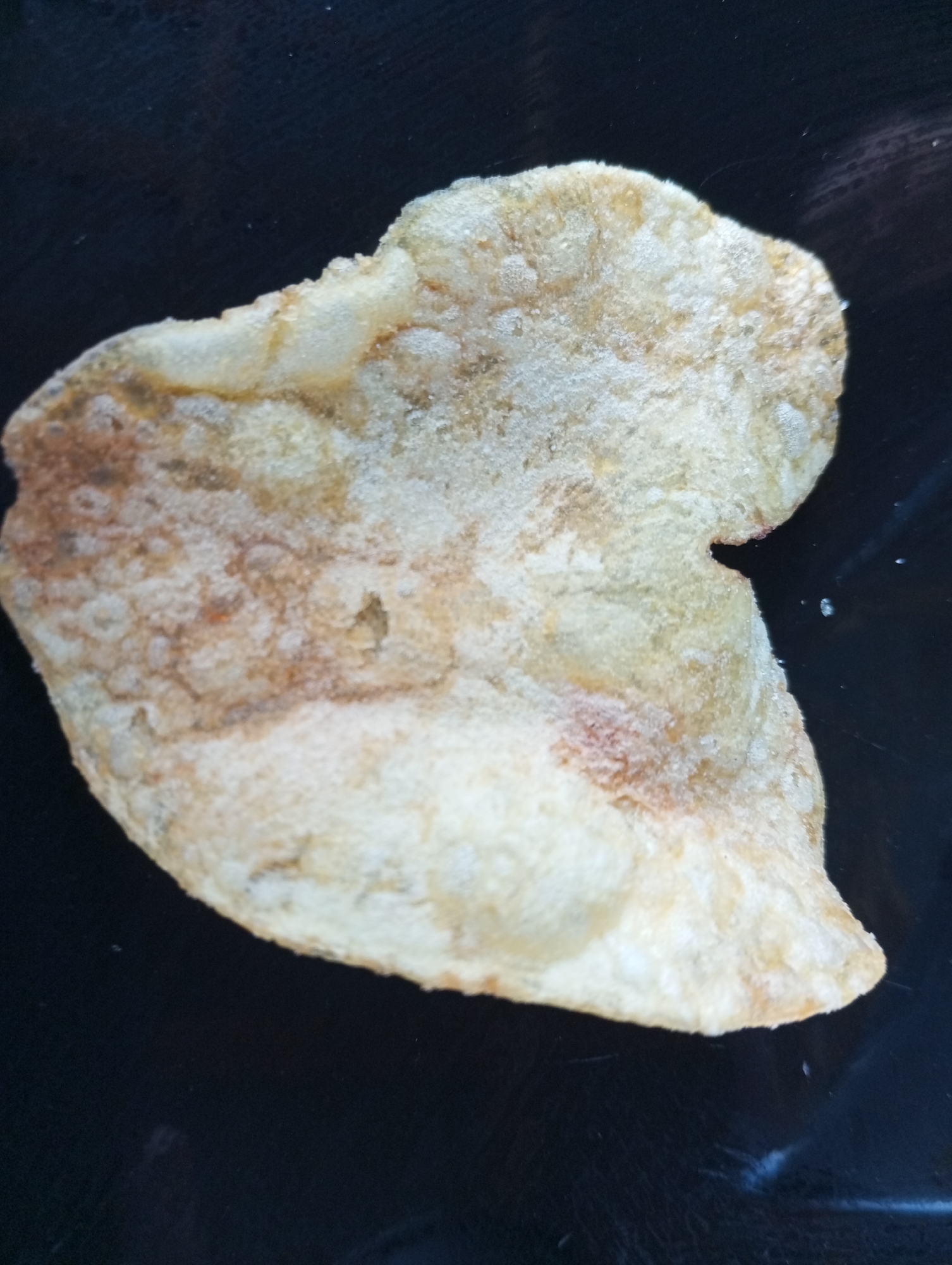 A single, irregular-shaped potato chip rests on a dark, reflective surface, possibly a plate or countertop. The chip is light brown and golden in color, with some darker, almost reddish-brown patches. Its surface appears slightly bumpy and textured, with a scattering of white powdery residue, suggestive of salt or seasoning. The background is uniformly dark and out of focus. There is no indication of any specific location within the city of Dymer, Ukraine, or any people, actions, or interactions.