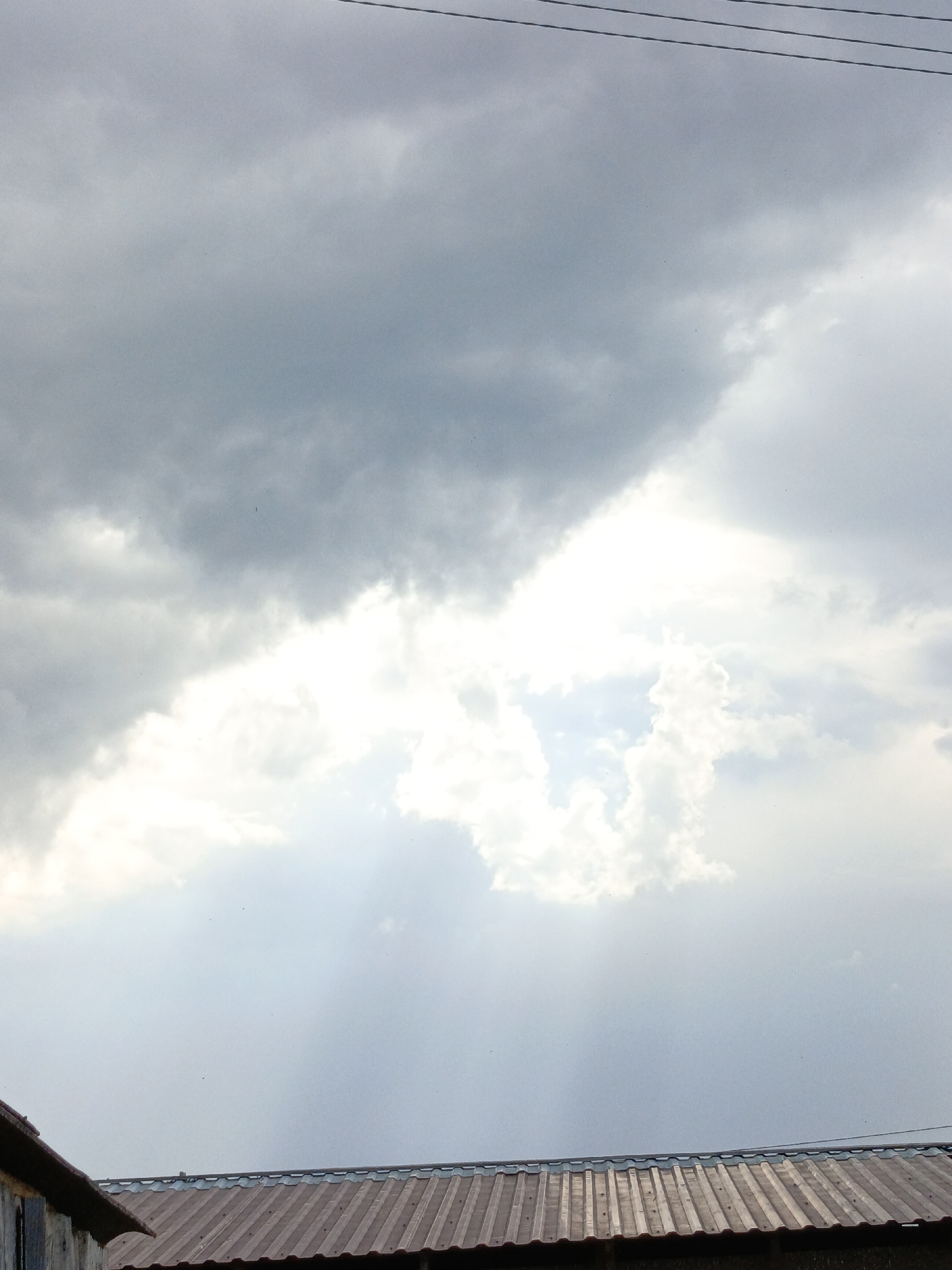 The image shows a cloudy sky over what appears to be a corrugated metal roof. The clouds are mostly gray, with a bright area where the sun is breaking through. The location is likely Zalesovo, Russia, as requested, but without specific landmarks, it is impossible to confirm. The scene depicts an overcast day with a potential break in the clouds. Power lines are visible in the upper part of the image.