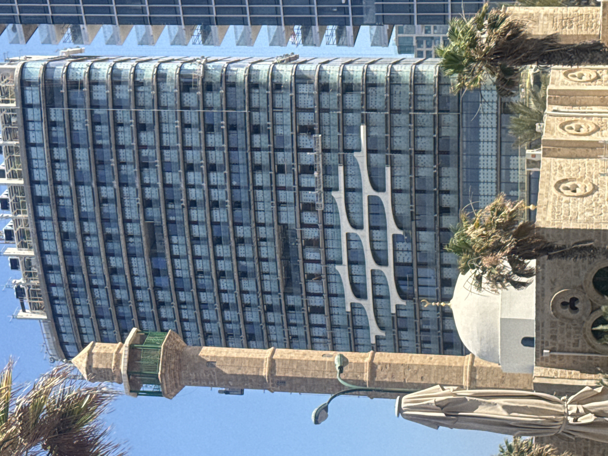 The image captures a juxtaposition of old and new architecture in Tel Aviv, Israel. In the foreground, a stone minaret and a white dome of a mosque stand against a bright blue sky. To the left of the minaret, a beige umbrella, covered with a tarp, is visible. A street lamp with two lights is also present on the left side. In the background, a modern, multi-story glass building dominates the scene, its facade reflecting the sky. The building appears to be under construction, with scaffolding and visible construction elements. Palm trees are scattered throughout the scene, adding to the tropical ambiance. The overall impression is one of historical religious structures existing alongside contemporary urban development.