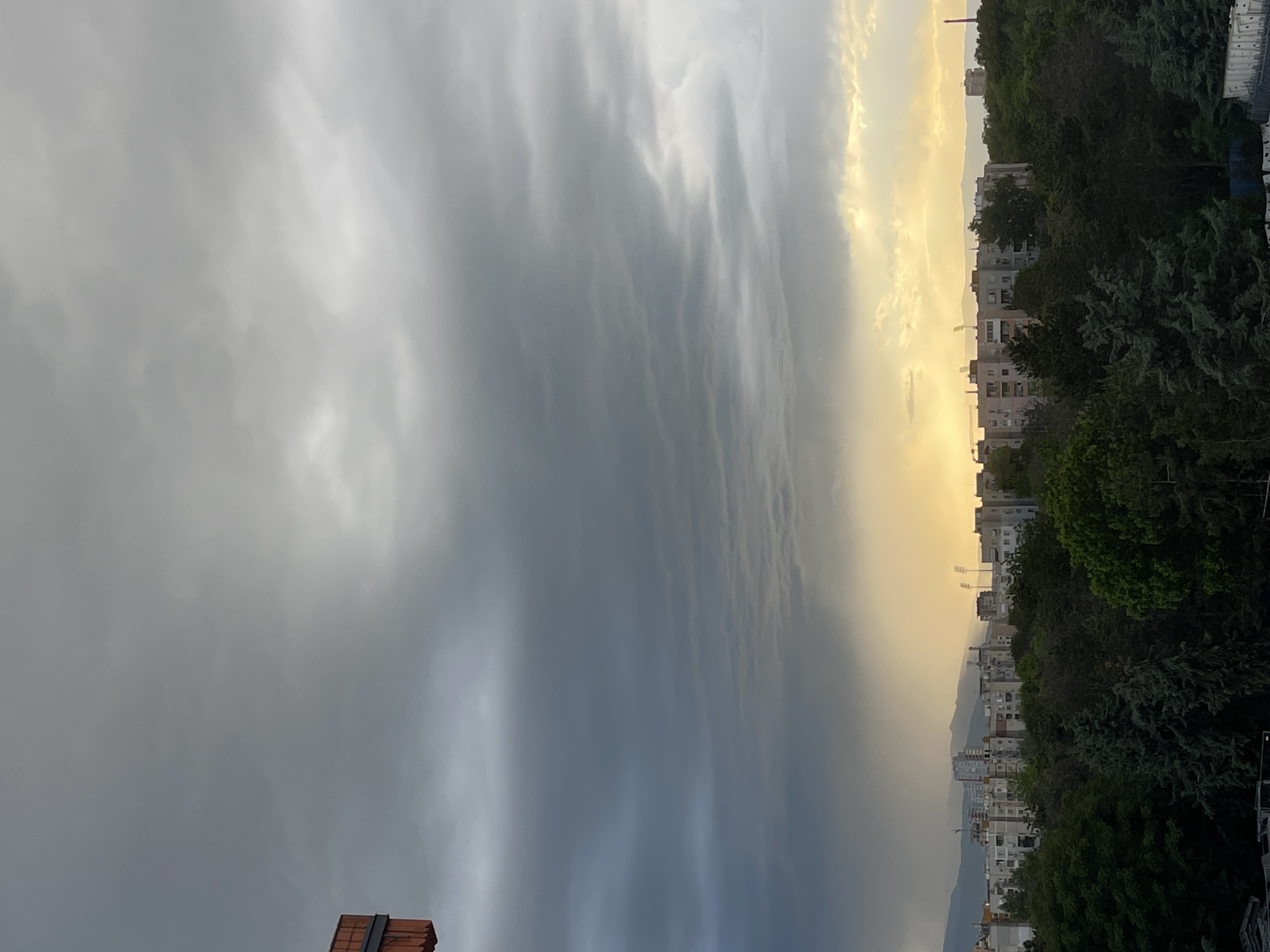 The image shows a view of Plovdiv, Bulgaria, from an elevated position, possibly a rooftop. The sky is divided between a dark, overcast portion and a brighter, sunlit area suggesting sunset or sunrise. The city skyline is visible, featuring apartment buildings nestled among trees. In the distance, mountains can be faintly seen through a golden haze. No people are visible in the scene.
